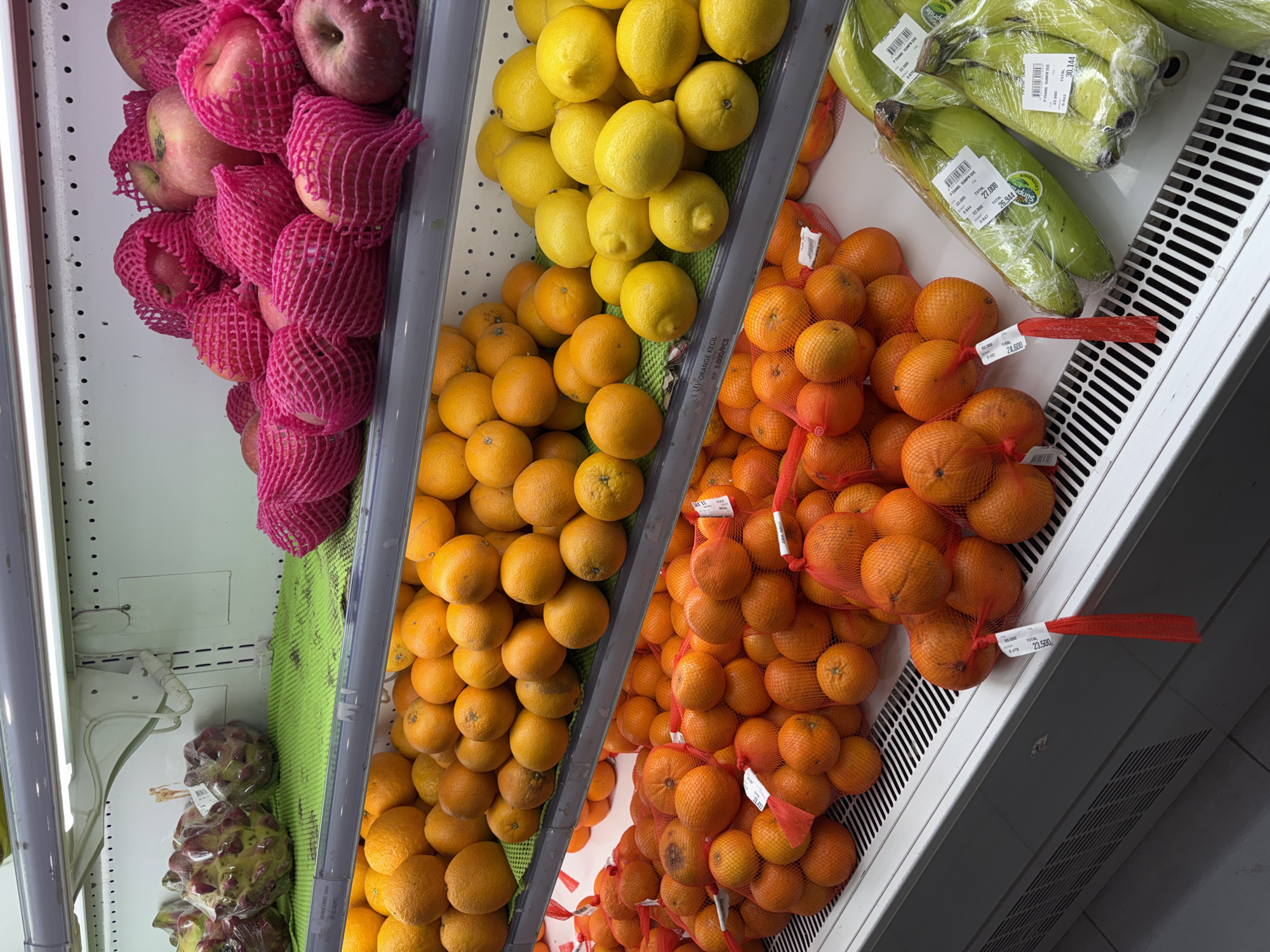 In a refrigerated supermarket display case, rows of fresh produce are neatly arranged on white shelves. The top shelf features several large, spiky green fruits wrapped in clear plastic. To their right, a pile of red apples, each individually encased in a bright pink mesh netting, creates a vibrant contrast. Below this, the second shelf is laden with oranges. On the third shelf, more oranges are displayed, some loose and others in red mesh bags. The bottom shelf showcases bright yellow lemons and green bananas, also packaged in plastic wrap, with price tags visible. The environment appears to be a well-lit grocery store, and given the presence of specific Indonesian product labels such as "ORANGE KECIL" and "PISANG SUMPI IDE" with local currency pricing, it is highly likely located in Tanjung Uban, Indonesia.