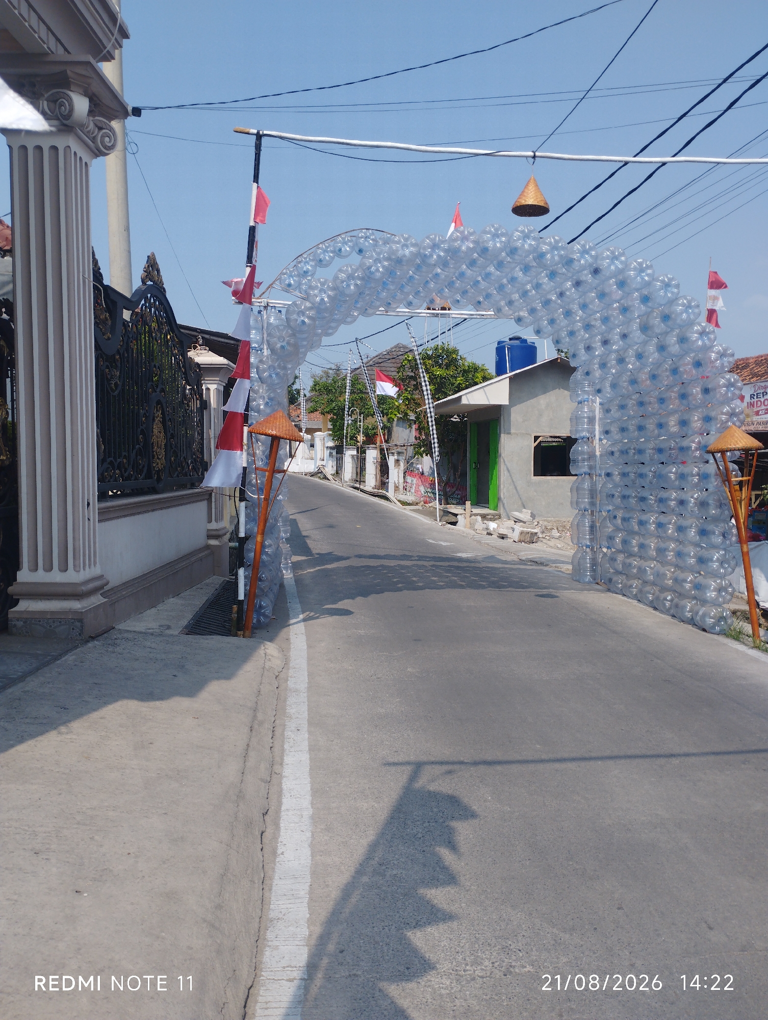 This is a medium shot, eye-level, outdoor photograph taken in Karawang, Indonesia. The image depicts a street adorned with an archway constructed from clear plastic water bottles. The archway spans across a road, which appears to be a local street with residential buildings on either side. The road surface is asphalt with a white line marking the edge of the pavement.
On the left side of the road, there is a decorative concrete pillar and a black metal fence. Several Indonesian flags, red and white, are prominently displayed, attached to poles and integrated into the archway structure. Some of these flags are small, triangular pennants, while others are larger rectangular flags. There are also some woven conical hat-like decorations hanging from wires.
In the background, a modest building with a blue water tank on its roof is visible. The sky is clear and blue. The overall impression is of a festive or celebratory decoration along a street in a residential area. The bottom left corner of the image shows the text "REDMI NOTE 11," and the bottom right corner displays the date and time "21/08/2026 14:22."