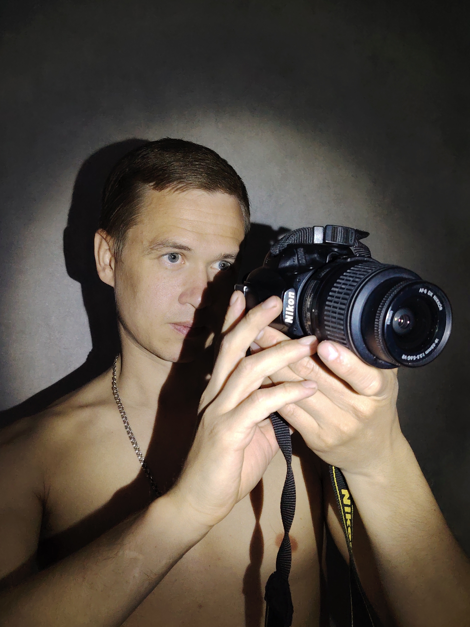 A shirtless man with short brown hair and blue eyes is holding a black Nikon camera in front of his face. He is wearing a silver chain necklace. The camera is angled slightly to the right, and the lens is visible. The camera strap is black with yellow "Nikon" lettering. The background is a dark, mottled gray. Shadows are cast on the man's face, chest, and the wall behind him. There is no evidence in the image to definitively suggest this photograph was taken in Chaikovsky, Russia.