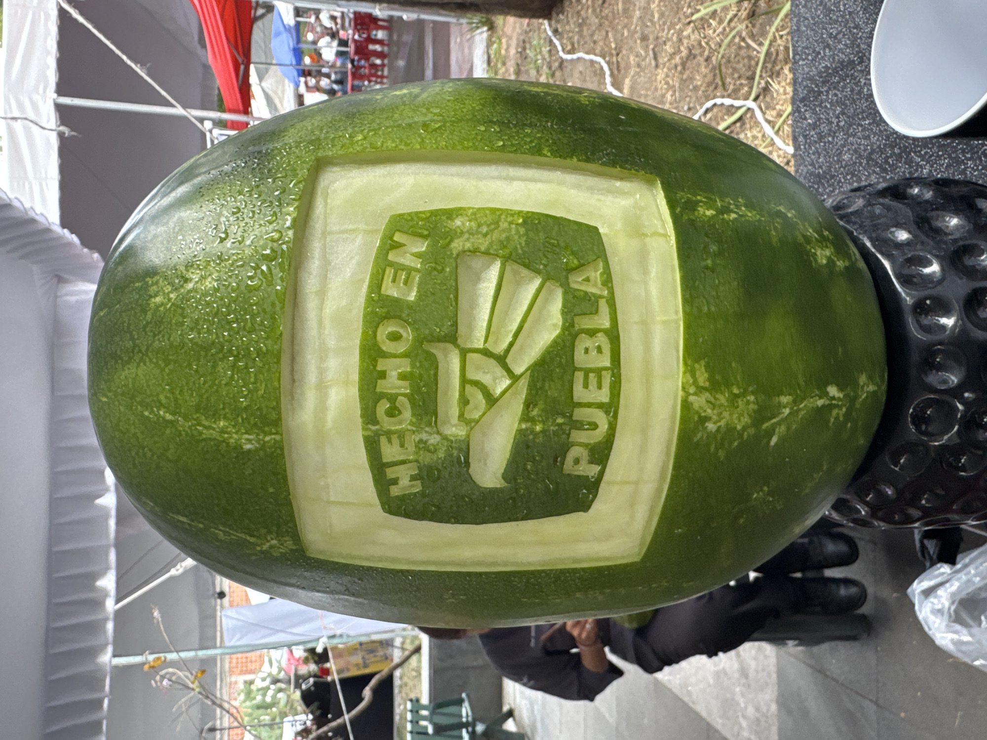 The image shows a watermelon with a carved design that includes the text "HECHO EN PUEBLA" (Made in Puebla) and a stylized icon, likely representing the state of Puebla, Mexico. The watermelon is green with some wet spots. It appears to be resting on a black textured surface and another round object. A white plate is visible in the upper-right corner. In the background, out of focus, there are tents, structures, and greenery, suggesting an outdoor setting. A person is visible in the lower part of the image wearing gloves. They appear to be holding or working on the watermelon.
