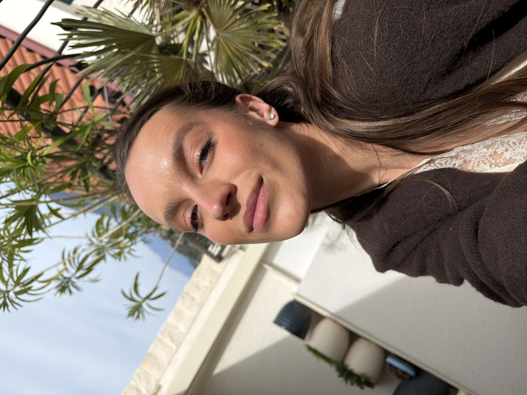 A woman is pictured in Trebesin, Montenegro, likely on a balcony or patio. The image, taken from a low angle and rotated to the side, shows her from the shoulders up. She is fair-skinned, has brown hair with highlights, and is wearing a brown top over a white, lace-trimmed garment. The woman has a stud earring in her left ear. The background includes foliage from a palm tree and other plants. White painted walls and several pots are also partially visible. In the distance, a building with a red-tiled roof and a hint of sky can be seen.