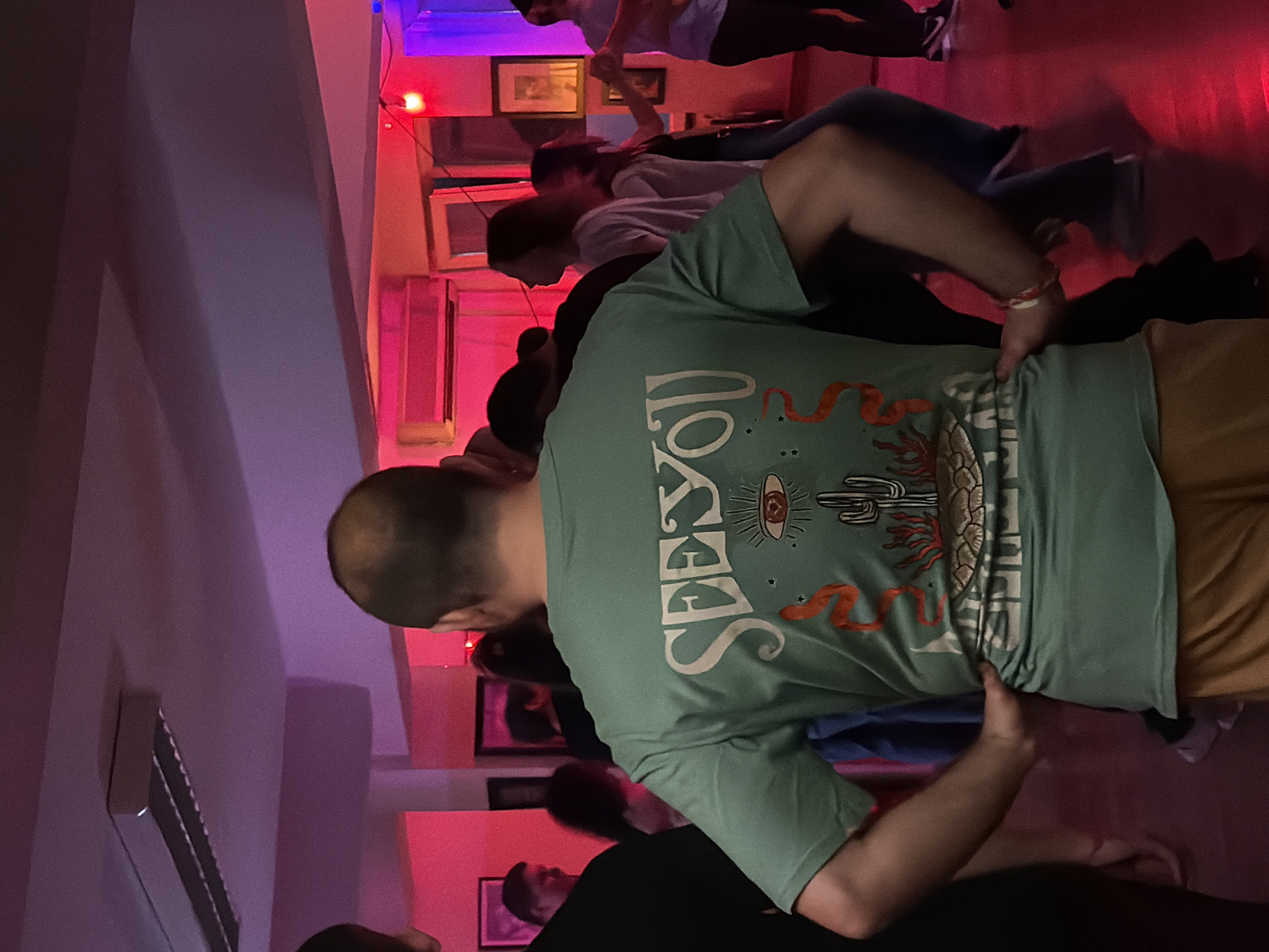 The image shows a gathering of people indoors. The main subject is a person wearing a light green t-shirt with the words "SEE YOU" printed on the back along with images of a cactus and an eye, wearing khaki pants. A group of people in the background are standing or walking. The room is lit with red and blue lights, and some framed pictures are on the wall. The location appears to be a club or bar in Sofia, Bulgaria.