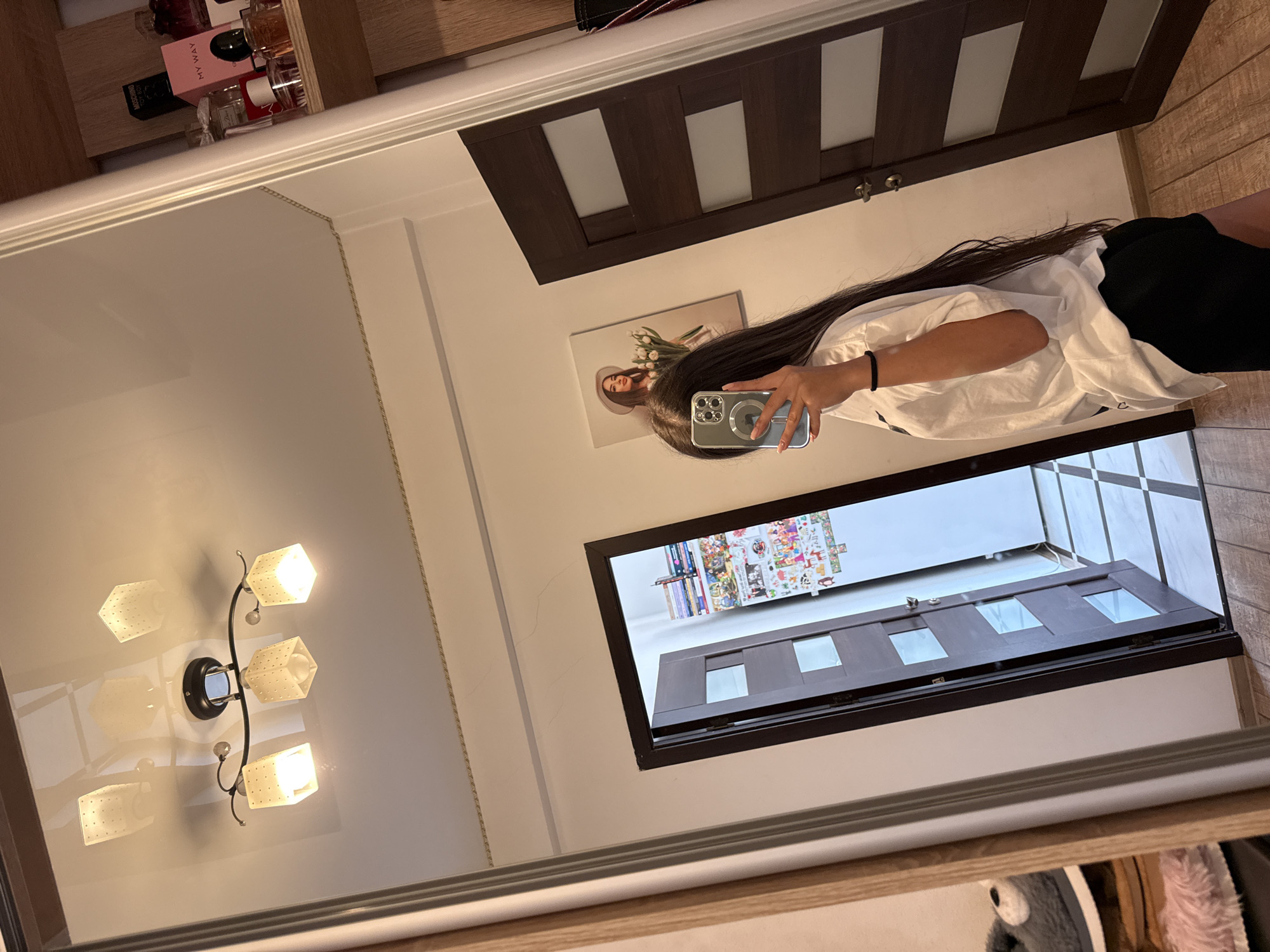 A young woman with long dark hair is standing in front of a large mirror, taking a selfie with her phone. She is wearing a white oversized t-shirt and black shorts. The mirror reflects the room she is in, which appears to be a hallway or entryway of an apartment in Chișinău, Moldova.
In the reflection, we can see a wooden door with glass panels, a light fixture on the ceiling, and a framed picture of a woman holding tulips on the wall. To the right, a shelf is visible with several perfume bottles. On the left side of the mirror, a glimpse of what appears to be a shoe rack with fluffy slippers can be seen. The floor is tiled with a checkered pattern in the background and wooden planks in the foreground. The overall impression is of a well-lit residential interior.