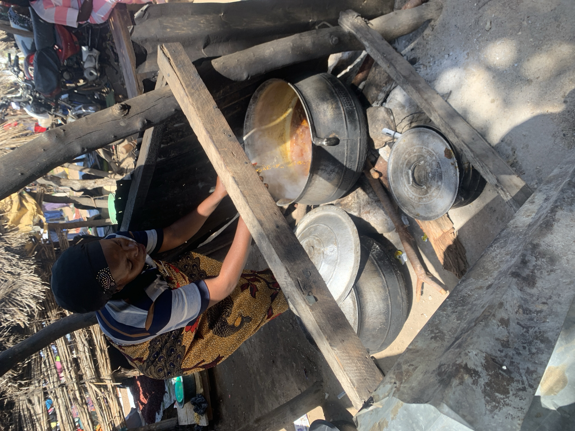 A woman, wearing a headscarf and a patterned dress, is tending to a pot of stew cooking over an open fire. The pot is large and black, and steam is rising from it. The woman is reaching towards the pot with her left hand.
The scene appears to be outdoors, possibly in a village or rural area, given the rustic construction of the cooking area with wooden beams and a dirt floor. There are other metal pots and lids scattered around, as well as some debris. In the background, a pile of motorcycles and other items suggest a busy or cluttered environment. The lighting suggests it is daytime, with bright sunlight casting shadows.
This image captures a moment of everyday life, highlighting the preparation of food in a traditional setting. The woman's posture and the presence of the cooking pot indicate an activity of sustenance. The overall impression is one of resourcefulness and resilience in a simple setting. There is no visible text in the image.