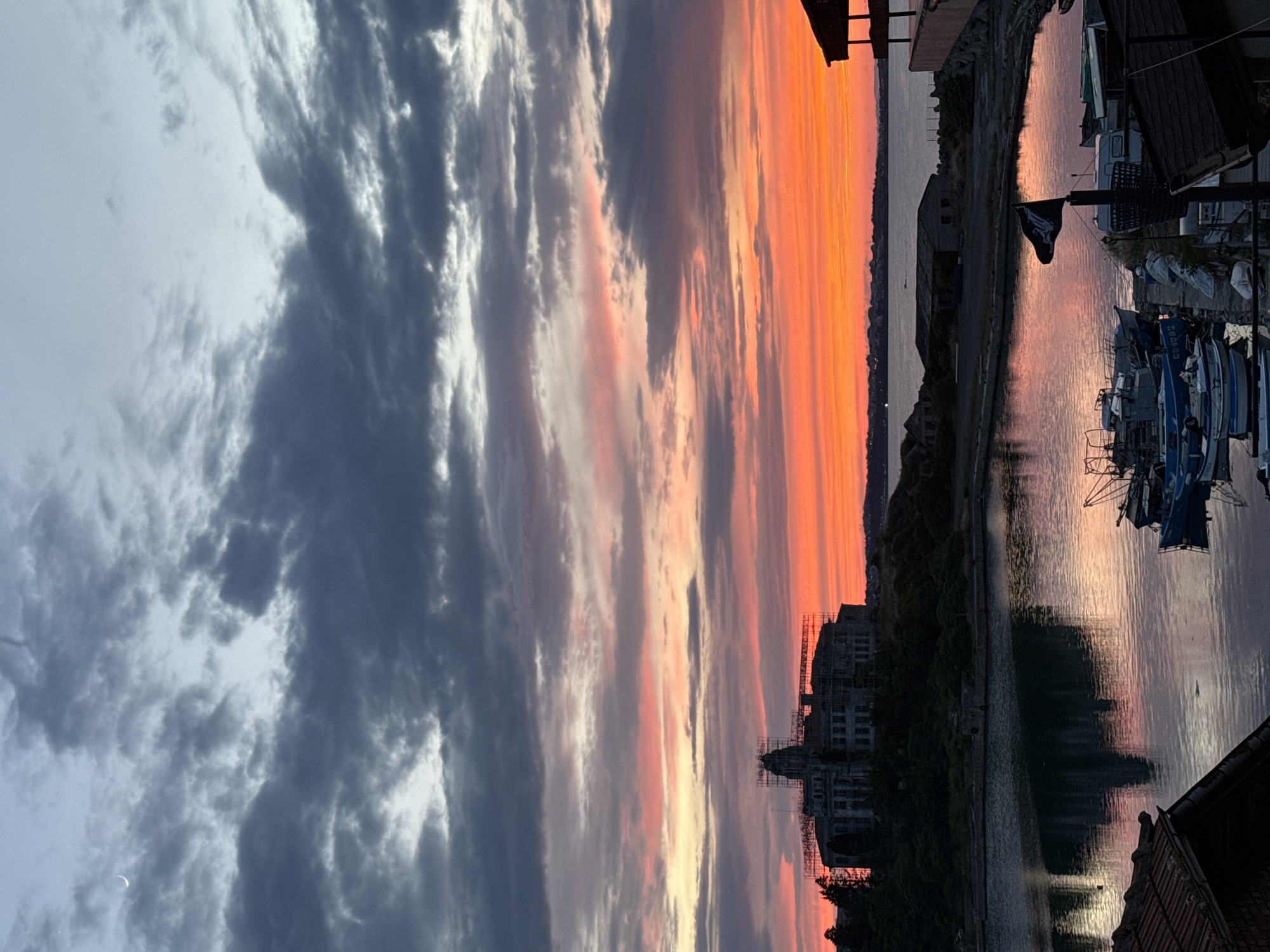 The image depicts a sunset over the bay in Sozopol, Bulgaria. A prominent, multi-story building, possibly a former school or public institution, is visible on a hill overlooking the water. The building appears to be under renovation, with scaffolding surrounding a central tower. The sky is filled with dramatic clouds, ranging from dark gray to vibrant hues of orange and pink, reflecting in the calm water of the bay. Several boats are moored in the foreground, some appearing to be small fishing vessels. A black flag with a skull and crossbones design is flying from a pole on the dock. The overall atmosphere is serene and picturesque, capturing a moment of twilight beauty in the coastal town.