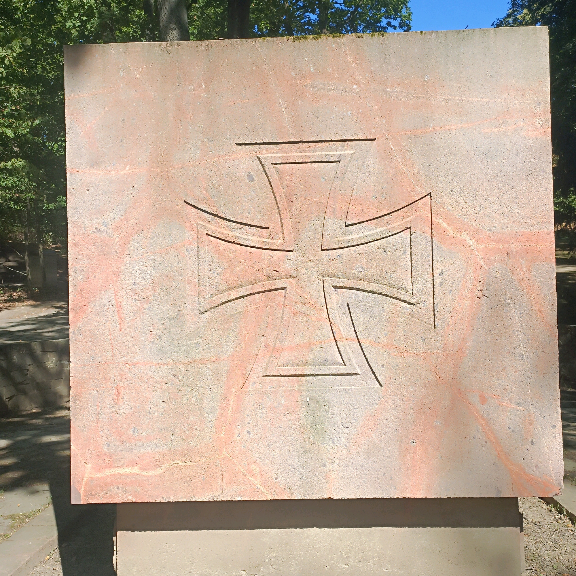 The image shows a large, square stone monument with an etched Iron Cross symbol in the center. The stone is a light reddish-pink color with grey speckling and some red staining. The Iron Cross is a stylized cross with flared ends, carved deeply into the stone. The monument is outdoors, with trees and foliage visible in the background under a clear blue sky. The monument appears to be part of a memorial or historical marker, given the symbol and its presentation. This monument is located at the Nordfriedhof (Northern Cemetery) in Wiesbaden, Germany, specifically at the Soviet War Memorial.