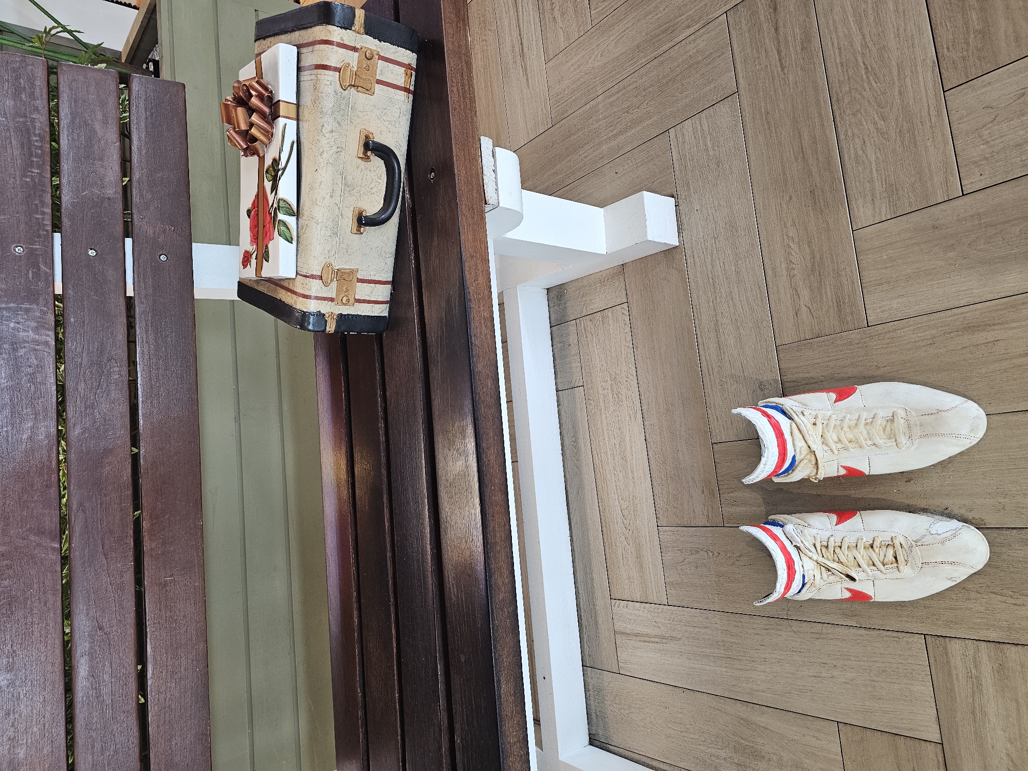 A pair of white athletic shoes with red and blue striped ankle socks are placed on a light brown, herringbone patterned floor. Behind the shoes, a dark brown wooden bench is visible, with a vintage-style suitcase and a gift-wrapped box on top of it. The suitcase is light-colored with red stripes and brass-colored latches and handle. The gift box is white and decorated with a red rose and a brown ribbon. The bench has white legs and is positioned outdoors, with greenery visible in the background. This scene is located in Denpasar, Indonesia.
