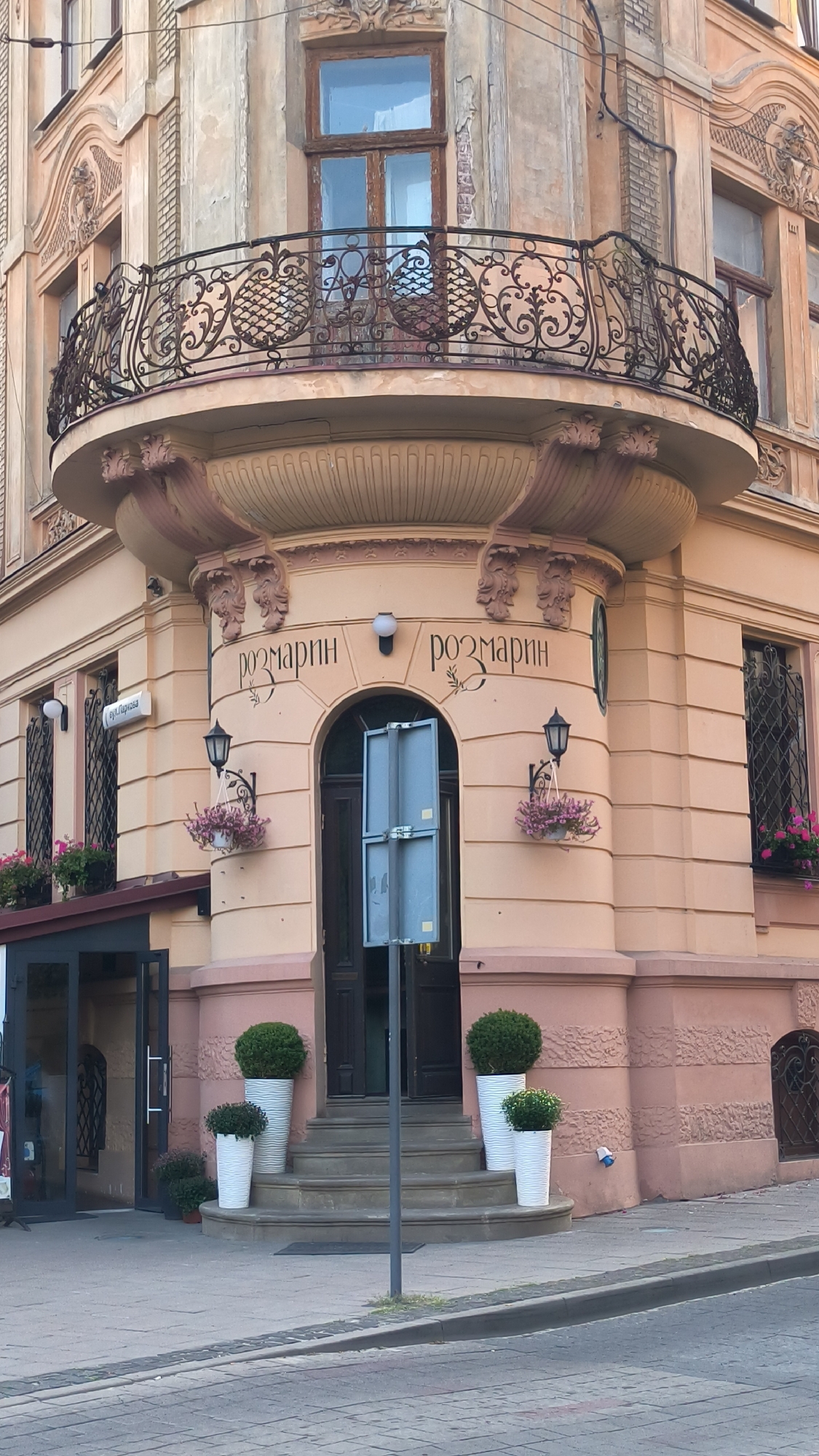 The image shows the corner facade of a building in Lviv, Ukraine, likely a commercial establishment. The building has a peach-colored stucco exterior with decorative architectural elements, including a curved balcony with ornate ironwork on the upper level. Below the balcony, the facade features two signs with the Cyrillic word "РОЗМАРИН" (Rozmarin), suggesting it's a business with that name, possibly a cafe or shop. The entrance is a dark wooden door accessed by a wide set of stone stairs, flanked by potted green plants in white planters. To the left of the entrance, a sign with Cyrillic script is visible on the building. Potted flowers hang from sconces near the signs. The street in front is paved with cobblestones. The overall impression is of an older, well-maintained European building.
