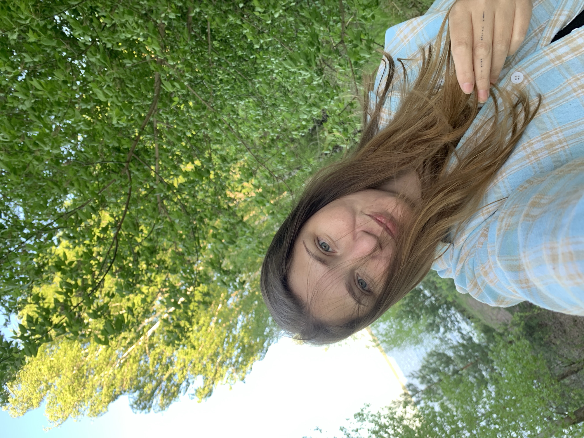 A woman with long, brown hair is taking a selfie outdoors. She is wearing a light blue and tan plaid shirt. Her hand rests on her chest near the collar of the shirt. The background shows green trees and a bright sky. The location appears to be a park or natural area in Chaikovsky, Russia. A body of water is visible in the distance.