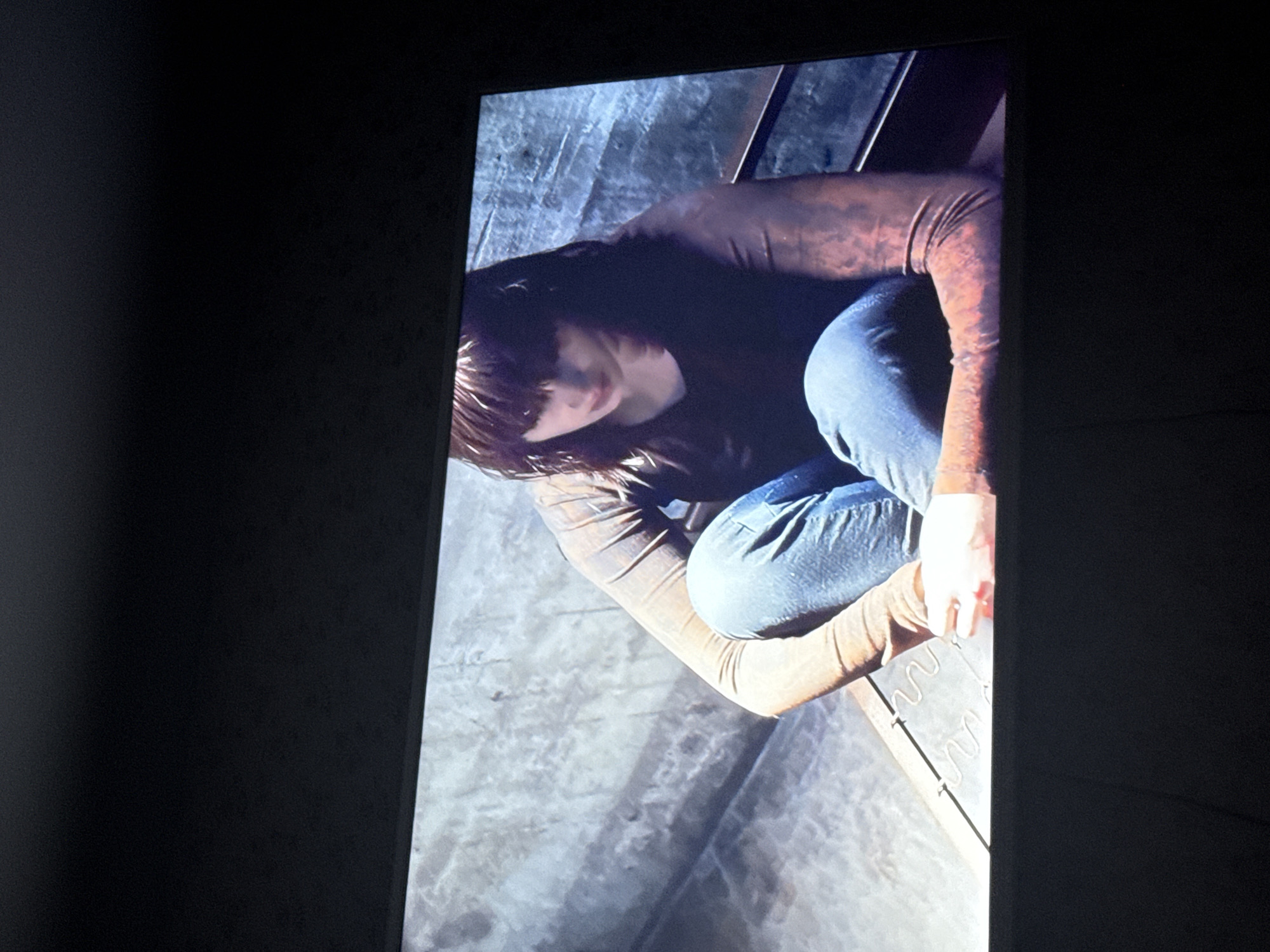A woman with dark, shoulder-length hair and bangs is crouching on what appears to be a concrete surface with metal bars or springs visible. She is wearing blue jeans and a dark, long-sleeved shirt with possible reddish-brown details on the arms. The lighting is dim, with a mix of shadows and highlights across the scene. The location is not identifiable as Mytishchi, Russia based on the visual information.