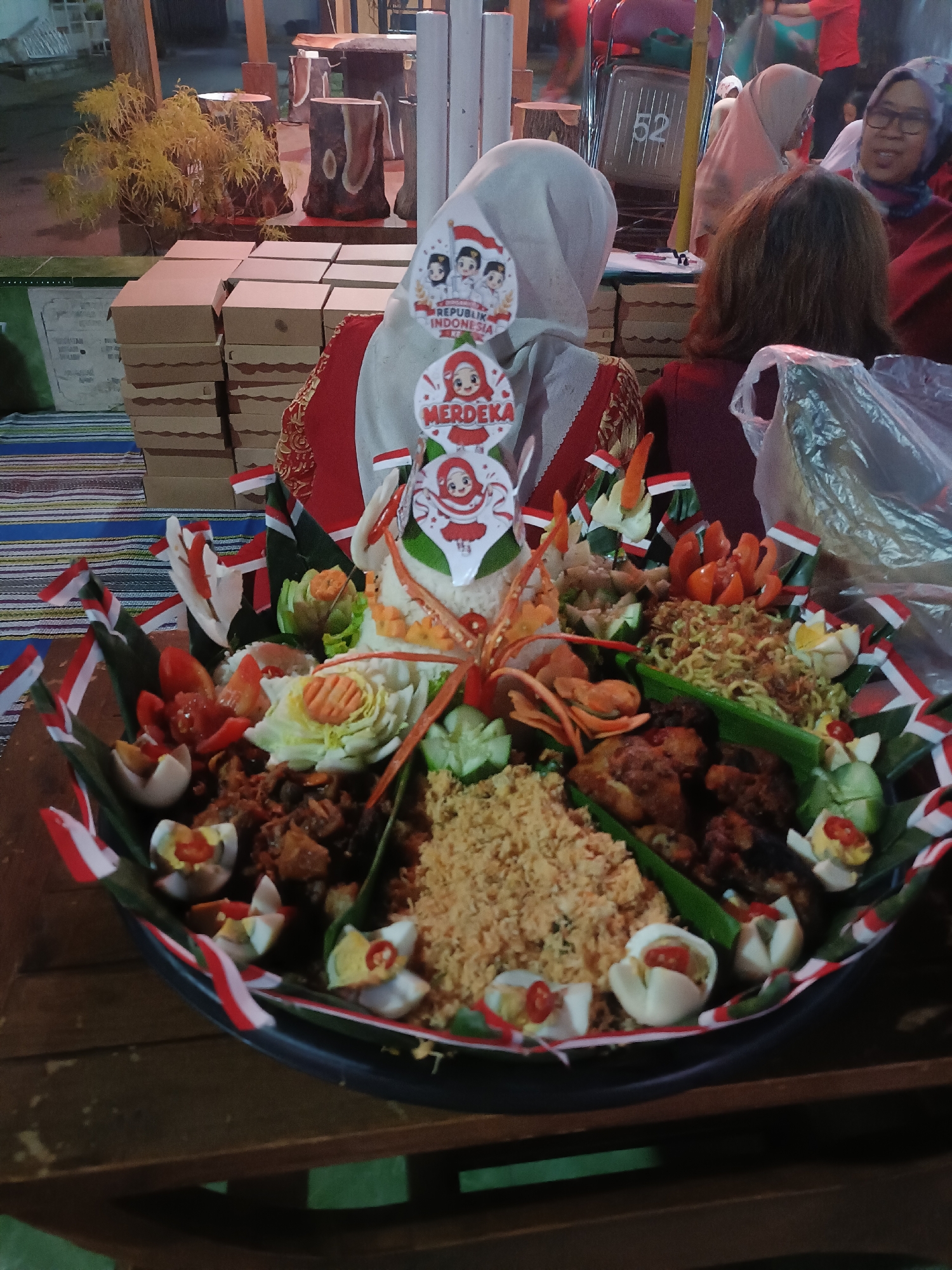This is a medium-shot, eye-level, outdoor photograph of a food offering, likely in Kediri City, Indonesia, given the Indonesian flags and "Merdeka" (Independence) decoration on a conical rice arrangement. The primary subject is a large, decorative platter of food, arranged in a conical shape using banana leaves as a base. The food includes rice, fried chicken, noodles, hard-boiled eggs, and various vegetables. Small Indonesian flags are placed around the perimeter of the platter. In the background, several people are visible, some wearing headscarves and traditional clothing. Boxes are stacked behind the food platter, suggesting a catering event or market stall. The overall lighting is dim, indicating it is likely evening.