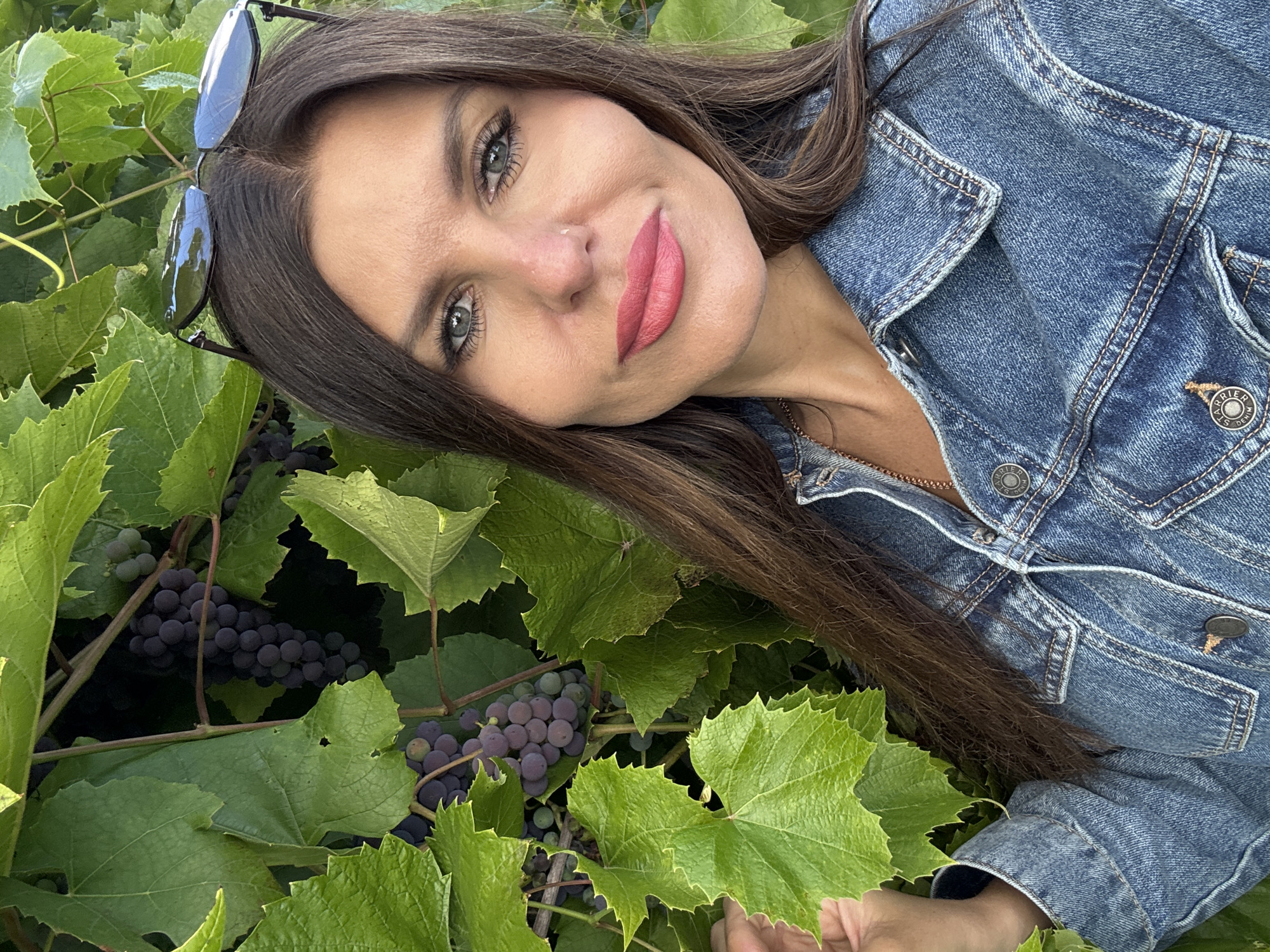 A woman with long brown hair, wearing a denim jacket and sunglasses perched on her head, poses for a selfie outdoors. She has on pink lipstick and her gaze is directed at the camera. Behind her, a dense canopy of green grape leaves and dark purple grapes frames the scene. The image appears to have been taken during daylight. The specific location in Dubovaya, Russia, is not discernible from the visual information provided.