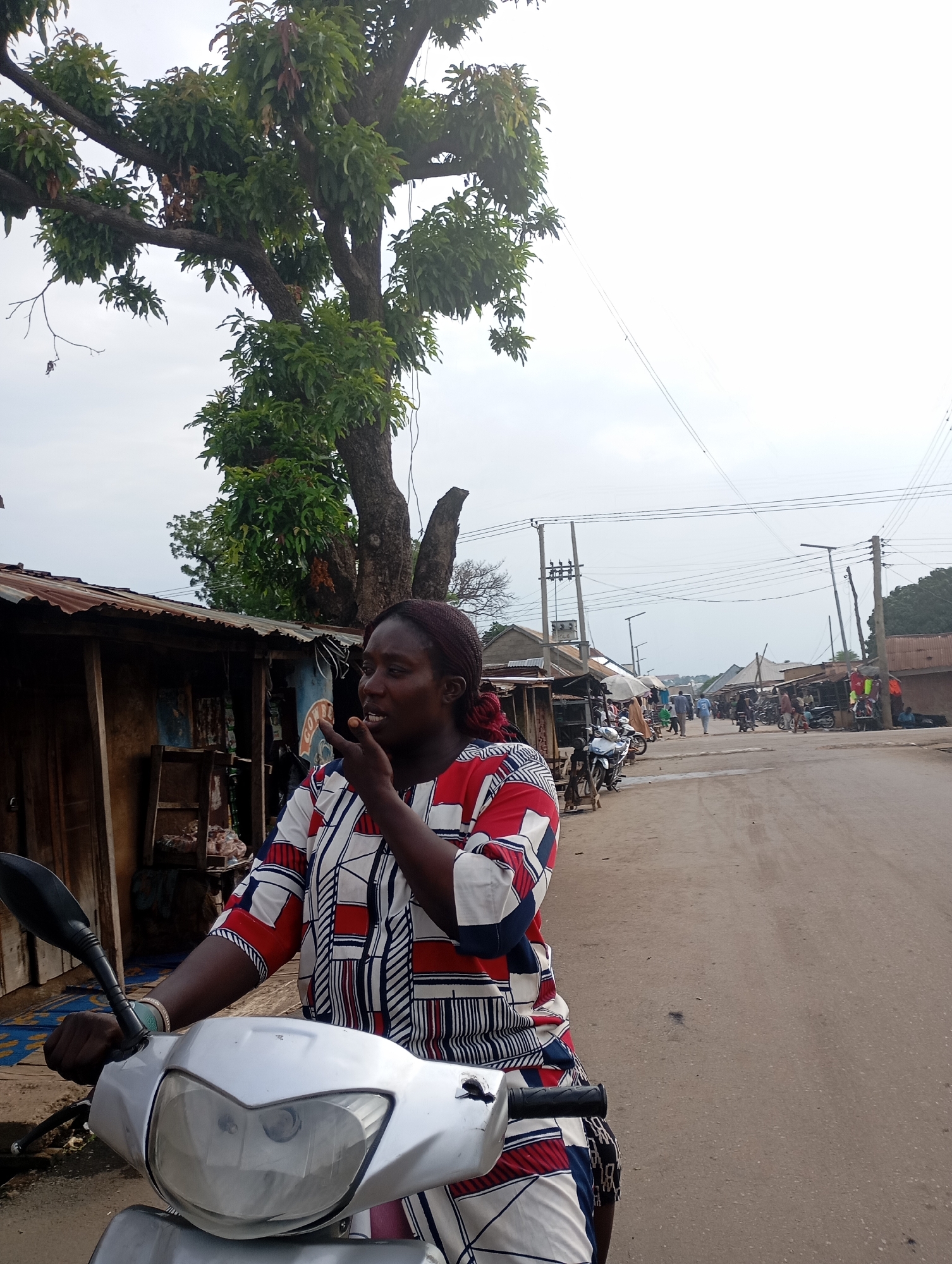 A woman is seated on a silver scooter in what appears to be a market street in Shendam, Nigeria. She is wearing a red, white, and blue patterned shirt and has her right hand raised to her mouth, with her index finger touching her lips. The scooter's handlebars and front panel are visible in the foreground. Behind her, a tree with green foliage stands to the left. Along the street, various stalls with corrugated metal roofs are set up, displaying goods and a few other motorcycles are parked. In the distance, more market stalls and people can be seen, with utility poles and power lines stretching across the sky, which is overcast. The road is paved with asphalt.