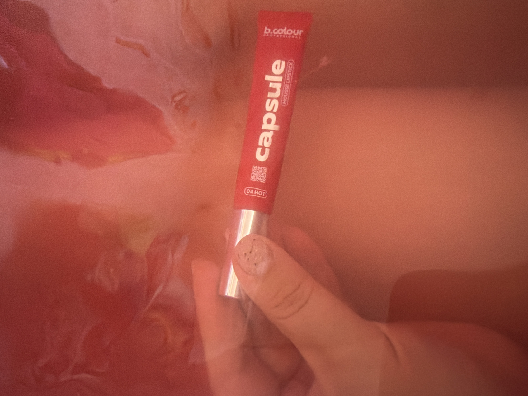 A hand holds a red tube of "b.colour professional capsule mousse lipstick 04 HOT" against a blurry, reddish-pink background that suggests water or a liquid surface. The location and city are unknown.