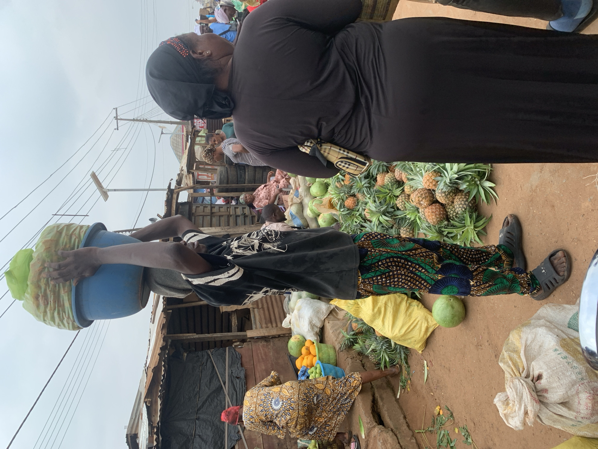 In Shagamu, Nigeria, a bustling open-air market is depicted. A young man, wearing a black t-shirt and patterned trousers, carries a large blue bucket filled with produce atop his head. The bucket is also topped with a large amount of green material. To his left, a woman in a yellow and brown patterned dress walks away, carrying a red headscarf. In front of her, a blue basket overflows with oranges, and various green fruits are visible. Piles of pineapples are scattered on the ground around the vendor stalls. A woman wearing a black headscarf and a long black dress stands to the right of the young man, facing away from the camera. The market appears to be set up along a dirt road, with makeshift stalls constructed from corrugated metal and wood. Overhead, power lines crisscross the sky. The overall scene suggests a typical day at a Nigerian local market.