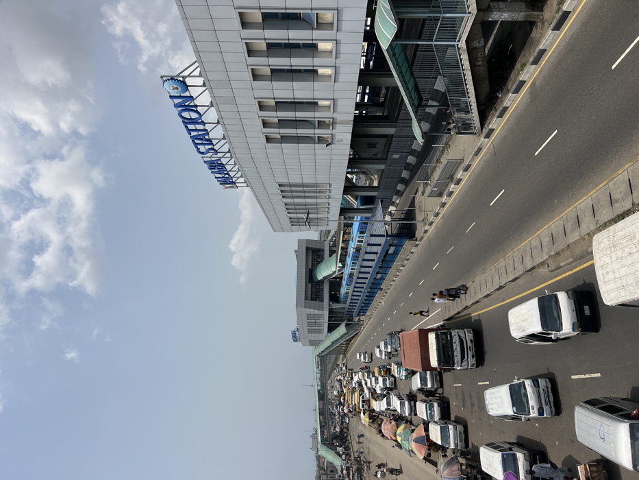 The image presents an elevated view of Alaba Station in Alaiyabiagba, Nigeria, under a bright, partly cloudy sky. The prominent Alaba Station building, a multi-level modern grey structure with "ALABA STATION" in large blue lettering on its roof, is situated on the right, featuring numerous rectangular windows and an elevated platform. A blue and white multi-car train is visible stationary on the elevated tracks at the station. Elevated pedestrian walkways and bridges, one with a green canopy, connect to the station and span across the primary road. Below, a multi-lane highway or major road extends through the scene. The left side of the road exhibits heavy traffic congestion, comprising numerous white vans, cars, motorcycles, and larger trucks, many appearing stationary. Alongside this congested section, multiple market stalls with various colored umbrellas (green, white, striped) are set up. Numerous individuals are seen walking among the vehicles and market stalls, indicative of commercial activity. On the right side of the main road, adjacent to the station, traffic is lighter, with a few groups of people standing on the sidewalk near a stairwell leading to a pedestrian bridge. At least 20 individuals are identifiable across the scene, engaged in transit and market-related movements.