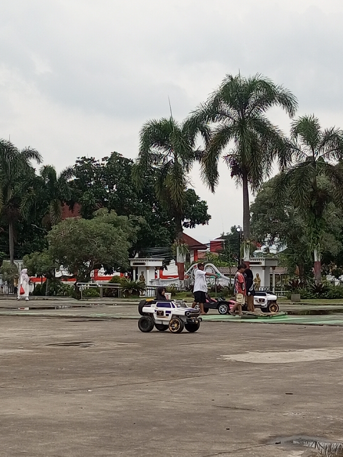 A partly cloudy day reveals a spacious plaza, where people congregate. A child drives a white toy monster truck in the foreground. Behind, two other children are near similar vehicles. Adults stand nearby, some watching the children. The plaza features palm trees and a red and white building with statues. The atmosphere is casual and active, indicative of a public gathering place.