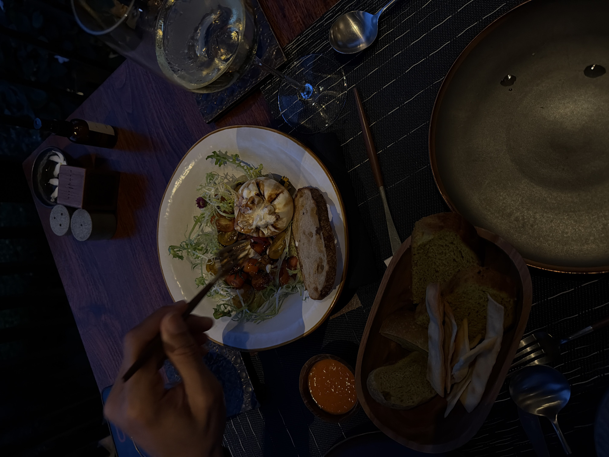 The image shows a dining table in a dimly lit setting, likely in a restaurant. The location is purportedly Kediri, Indonesia, though no elements in the image directly confirm this. On the table, there is a plate with what appears to be a salad featuring grilled garlic, tomatoes, and greens, alongside a slice of bread. Adjacent to the plate is a small bowl of orange sauce and a wooden bowl filled with various types of bread. There is a large, empty plate to the right. Cutlery, including a spoon and fork, is present. A wine glass containing a light-colored liquid, presumably wine, sits near the top left of the frame, along with a small bottle and what appear to be salt and pepper shakers. A person's hand is visible in the bottom left corner, holding a utensil (chopstick or similar). The table is covered with a black and white striped placemat. The environment suggests an upscale dining experience.