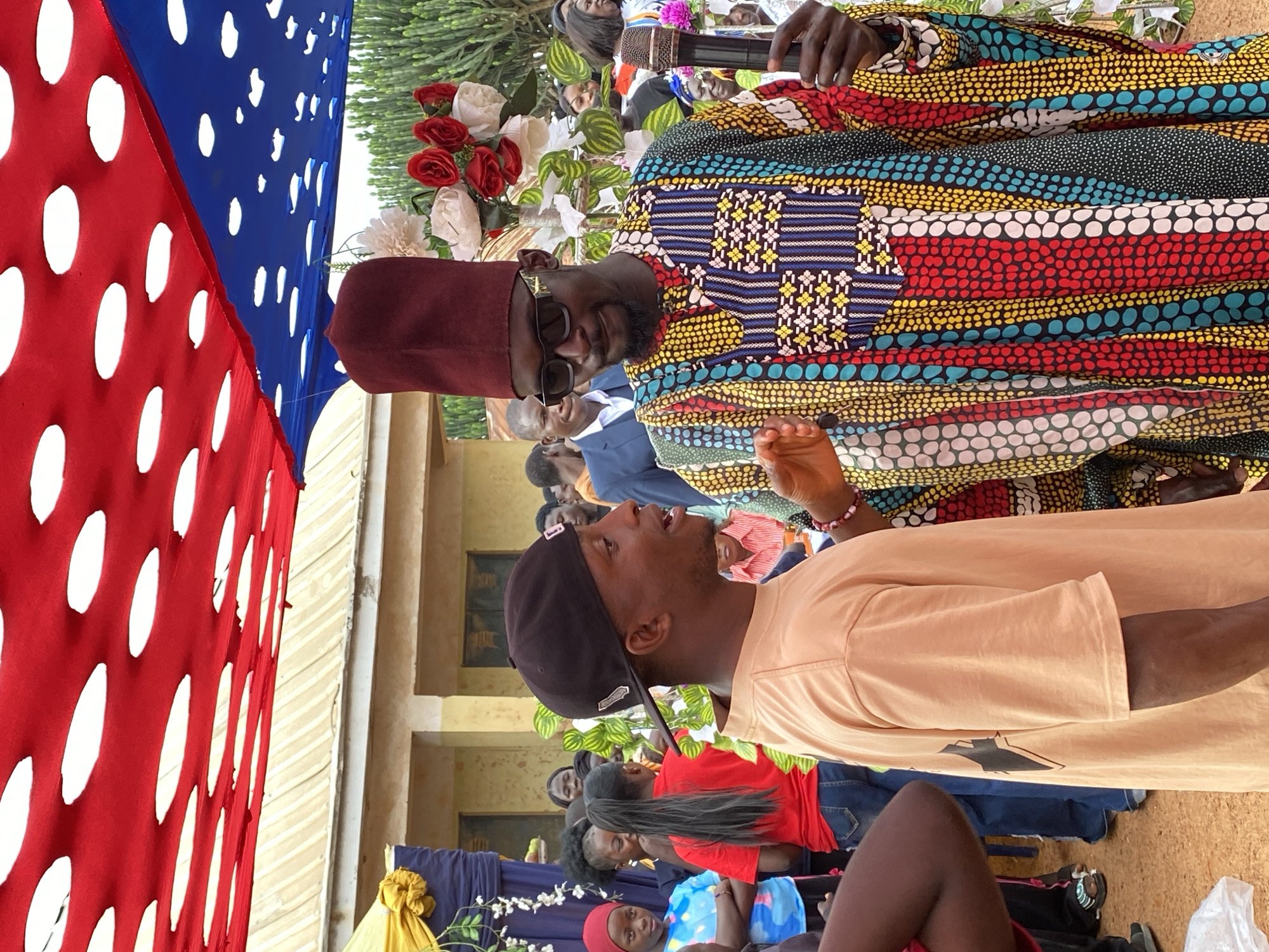 The image appears to be taken at an outdoor event in Kuru Dungus, Nigeria. A man in a colorful, patterned robe and a maroon hat is speaking into a microphone. He wears sunglasses and stands amidst floral decorations. A second man, wearing a black baseball cap and a light-colored shirt, looks up at the speaker. An audience is visible in the background. A red and blue decorative hanging with white circles is on the left side of the image. A building can be seen in the background. Attendees are seated in the foreground.
