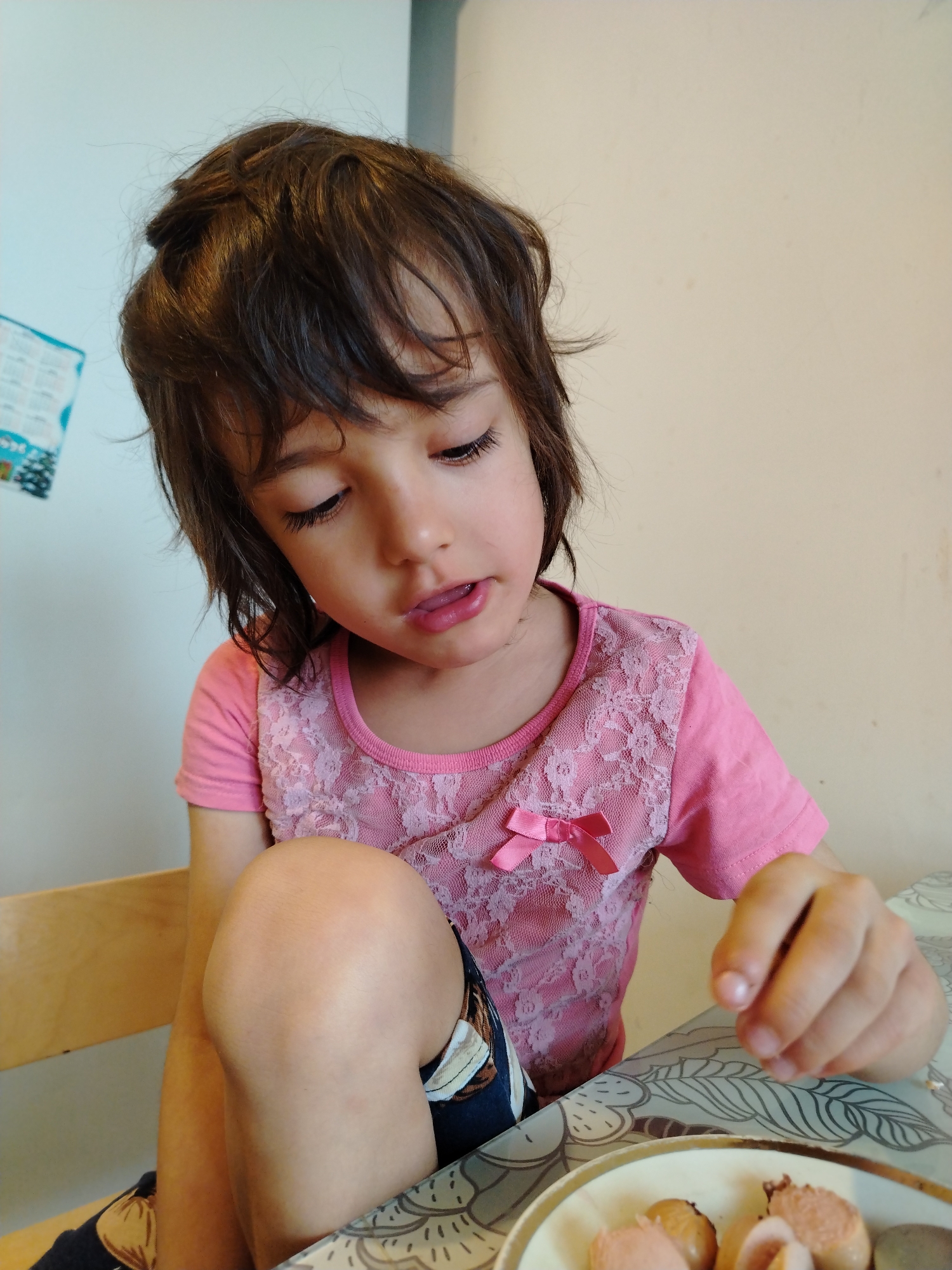 A young child with dark brown, disheveled hair is seated at a table, looking downwards at a plate of food. The child wears a pink short-sleeved top with a lighter pink lace overlay on the front, adorned with a small pink satin bow on the left chest. Patterned shorts or pants are visible on the child's lower body, with a bare knee prominently bent in the foreground. The child's left hand is raised slightly above a plate containing several cut pieces of sausage or similar cooked meat. The table surface features a grey and white patterned tablecloth. The background consists of a light blue wall on the left with a partially visible colorful poster, and a plain light beige wall on the right. A portion of a wooden chair is visible on the far left. The specific location in the city of Noviy, Russia, is not verifiable from the visual content of the image.