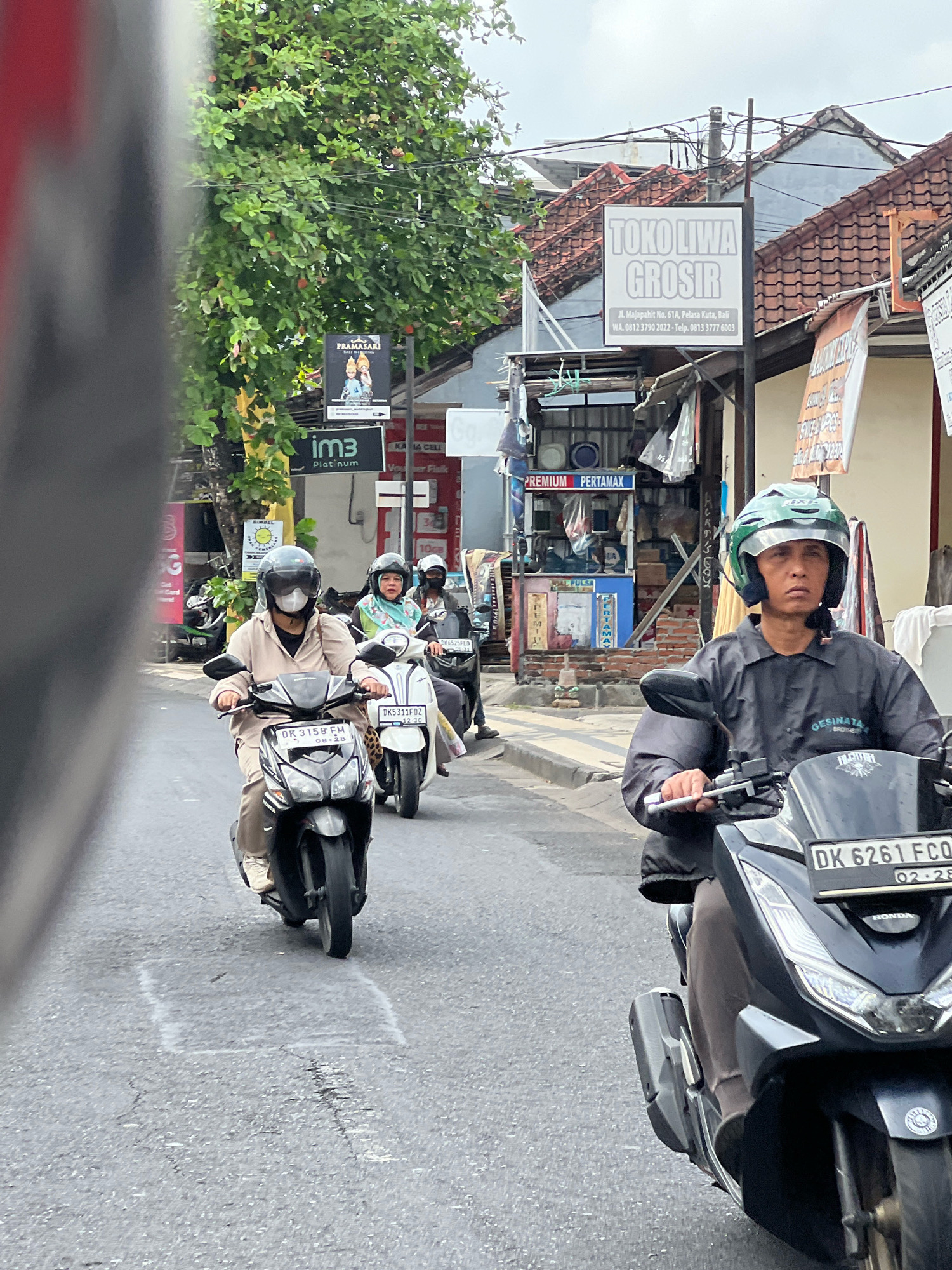 A motorcycle rider wearing a light brown long-sleeved shirt and tan pants drives a dark-colored scooter on a paved road in Kuta, Indonesia. They wear a white mask and a dark helmet with a clear face shield. The rider's scooter has a license plate that reads "DK 3158 FM". Behind this rider, another scooter driven by a person wearing a light blue hijab and a patterned scarf, is also visible. This scooter has a white body and a license plate reading "DK 625 FCD". The road is lined with buildings and shops, including one with a sign that says "TOKO LIWA GROSIR". Trees with green leaves are visible on the left side of the image. The overall scene depicts typical street traffic in a bustling urban environment.