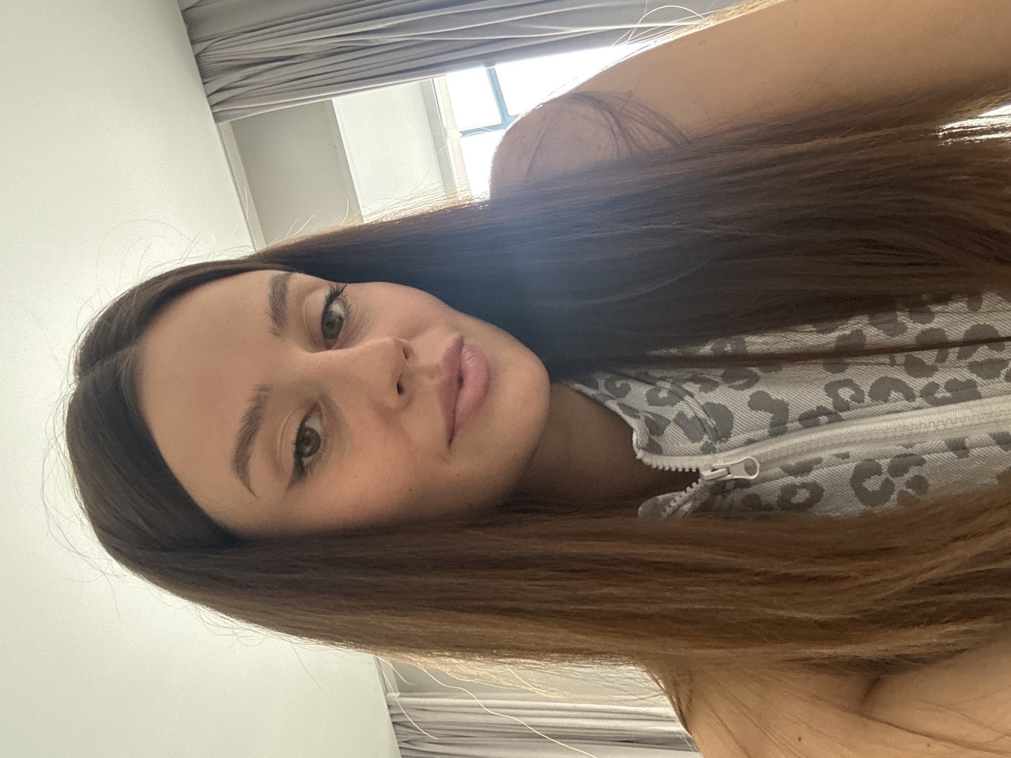 The image shows a close-up, upper-body portrait of a young woman with long, dark brown hair. She is wearing a zippered top with a grey and white leopard-print pattern. The background consists of a lightly-colored wall, grey curtains, and a window with bright daylight streaming through. The woman is looking directly at the camera with a neutral expression, her lips slightly parted. Her makeup includes winged eyeliner and lip gloss. The overall lighting suggests it is daytime, possibly indoors in a room with natural light. There is no visible information to suggest this image is located in Krasnoyarsk, Russia, or any specific city.