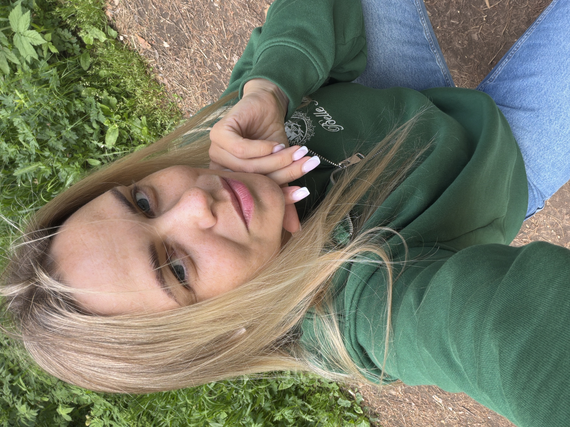 A woman is shown in a selfie-style, close-up image. She is wearing a green sweatshirt with an embroidered design and light-wash blue jeans. The woman has fair skin, blonde hair with dark roots, and light-colored eyes. Her fingernails are manicured with a white French tip design. The background includes green grass and a dirt path. The location cannot be identified as Votkinsk, Russia, based on the visual information provided.