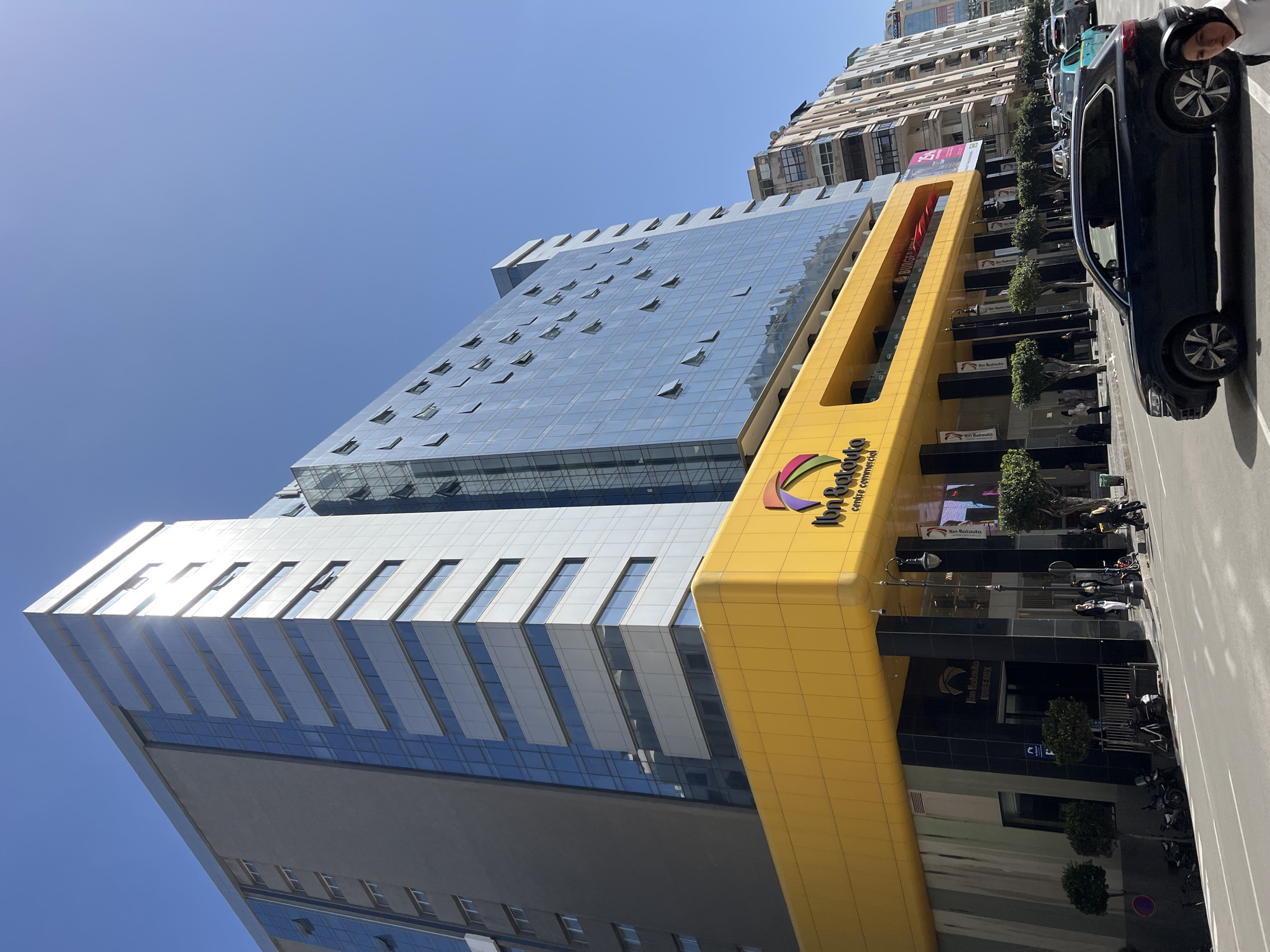 The image displays the Ibn Batouta Centre Commercial, a multi-story modern building located in Tanger, Morocco, under a clear blue sky. The building features a prominent, rounded yellow facade on its lower levels, with "Ibn Batouta centre commercial" and a multi-colored logo visible. Below the yellow section, the ground floor entrance areas are clad in black marble-like panels, with signs indicating "Ibn Batouta Bureaux."
Above the yellow base, the building's upper stories are constructed with a mix of light grey paneling and extensive blue-tinted glass curtain walls. Numerous windows on these upper floors are partially open, angled outwards. To the left, another section of the building exhibits darker grey and blue glass components. The sun reflects brightly off the building's surfaces.
On the street level, a wide sidewalk runs alongside a paved road. Several individuals are present on the sidewalk, some congregating near the main entrances of the commercial center, and others walking. A black Volkswagen Touareg SUV is in motion on the road in the foreground, with its driver visible through the window. Other vehicles are visible in the background traffic. Small, manicured trees line sections of the sidewalk, and several motorcycles are parked near the building. Streetlights and banners displaying the Ibn Batouta logo are also present. Signs for businesses such as "Zara" and "Burger King" are visible on the building's exterior. In the background to the right, multi-story residential and commercial buildings with varied architectural styles line the street.