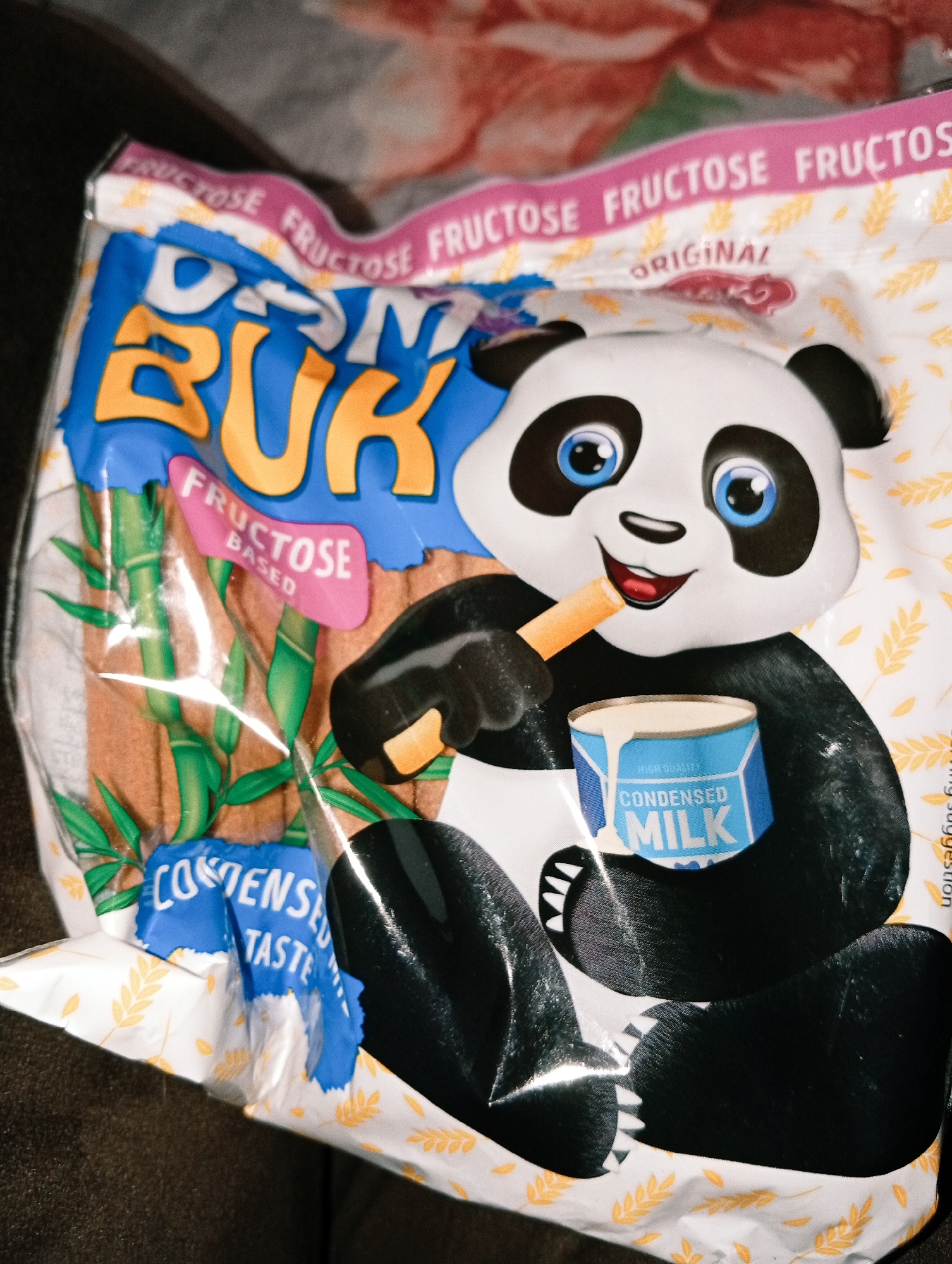 A close-up shot shows a partially crumpled bag of "Drim Buk" brand biscuit sticks. The bag is white with pink trim and depicts a cartoon panda holding a stick and a can of condensed milk. The panda has black ears, eyes, and paws, and blue irises. The biscuits are shown with green bamboo in the background. The words "Fructose" and "Condensed Taste" are printed on the bag. The location is not provided or discernible from the image.