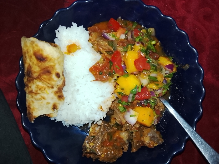 A blue plate holds a vibrant meal. White rice sits beside a folded, golden-brown flatbread. The main dish features braised meat topped with a colorful mango salsa, containing diced mango, red tomato, and red onion, garnished with cilantro. A sliver of a silver utensil peeks out from under the food. The plate rests on a reddish-burgundy surface.
