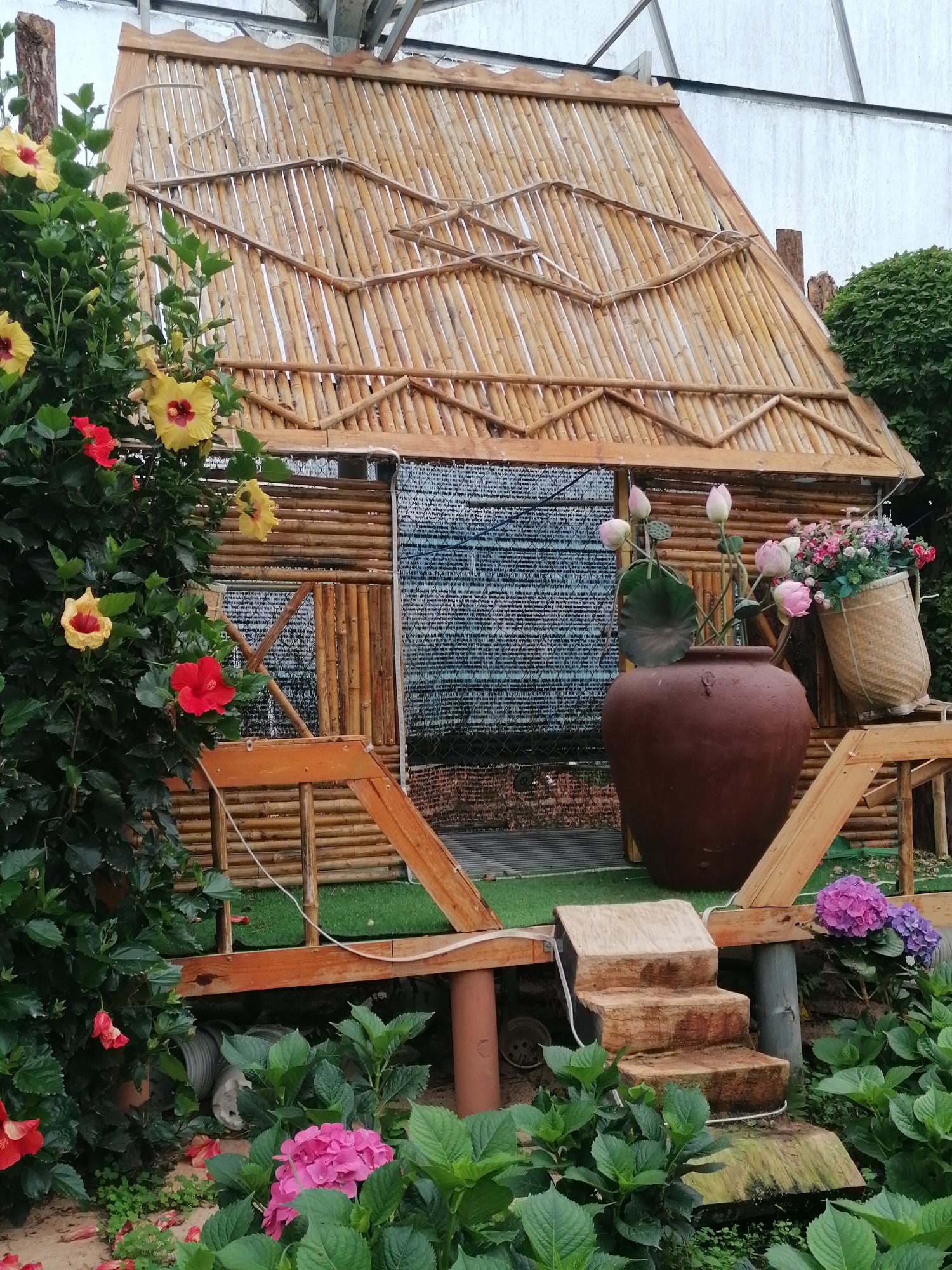 This image presents a serene and vibrantly decorated garden scene, likely set in Lạc Dương, Vietnam, given the cultural context of the architecture and flora. The central subject is a charming, rustic hut or structure, meticulously crafted from bamboo and natural wood. Its steeply pitched roof is a prominent feature, constructed from numerous bamboo poles laid side-by-side, with additional decorative bamboo patterns on its surface. The front of the hut also incorporates horizontal bamboo logs, partially covered by a dark, mesh-like screen or curtain, suggesting an enclosed space.
The hut stands on a raised wooden platform covered with artificial green turf, and three rustic steps, carved from solid logs, lead up to it from the ground. A simple wooden railing surrounds the front of the platform.
The scene is rich with lush plant life. On the left, a large, leafy bush is adorned with numerous hibiscus flowers in striking shades of bright yellow with deep red centers, and vibrant red. Towards the bottom of the frame, more red petals are scattered on the ground, and dense green foliage is visible, including vibrant pink and purple hydrangea bushes blooming below the hut's platform.
To the right of the hut's entrance, a large, rounded brown ceramic pot holds a beautiful arrangement of pink lotus buds, complete with their distinct green leaves and seed pods, adding an element of calm beauty. Next to this pot, a woven bamboo basket overflows with a colorful assortment of artificial flowers, predominantly in shades of pink, purple, and white.
The lighting suggests it is daytime, possibly overcast, contributing to the soft and even illumination of the scene. In the background, hints of a larger structure with metal beams are visible against a light sky, possibly indicating a greenhouse or a park setting. There are no people visible, and no discernible text appears in the image. The entire composition exudes a peaceful, natural, and inviting atmosphere.