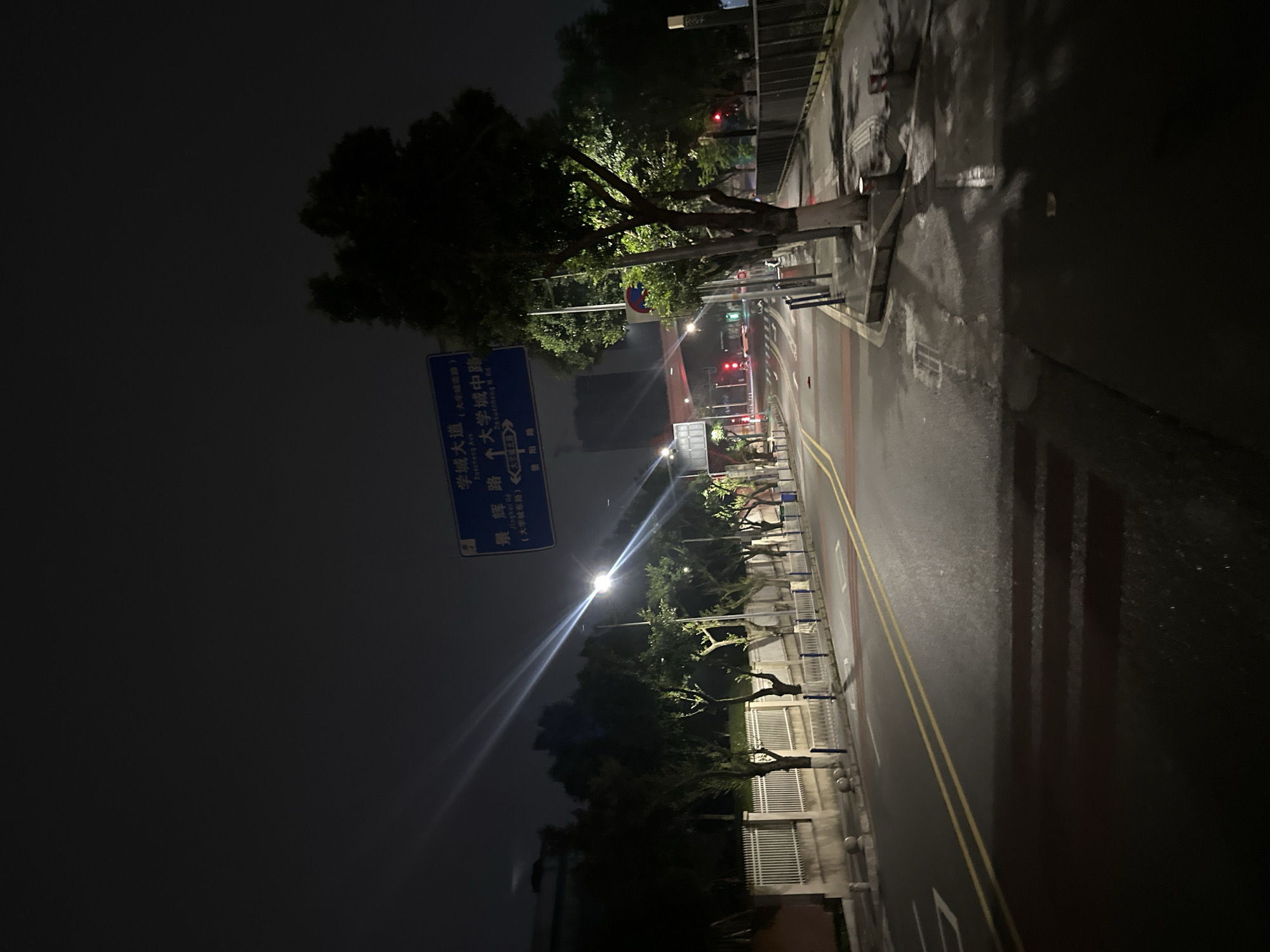 A street scene at night in the Chongqing High-tech Zone, China. A blue street sign indicates "Xuecheng Ave" (University Town South Rd) and "Jinghua Rd" (University Town Middle Rd). The road is illuminated by streetlights, casting long shadows. Trees line the sidewalk, and a white fence runs along the side of the road. The street appears wet, suggesting recent rain. No people or vehicles are visible in the image.