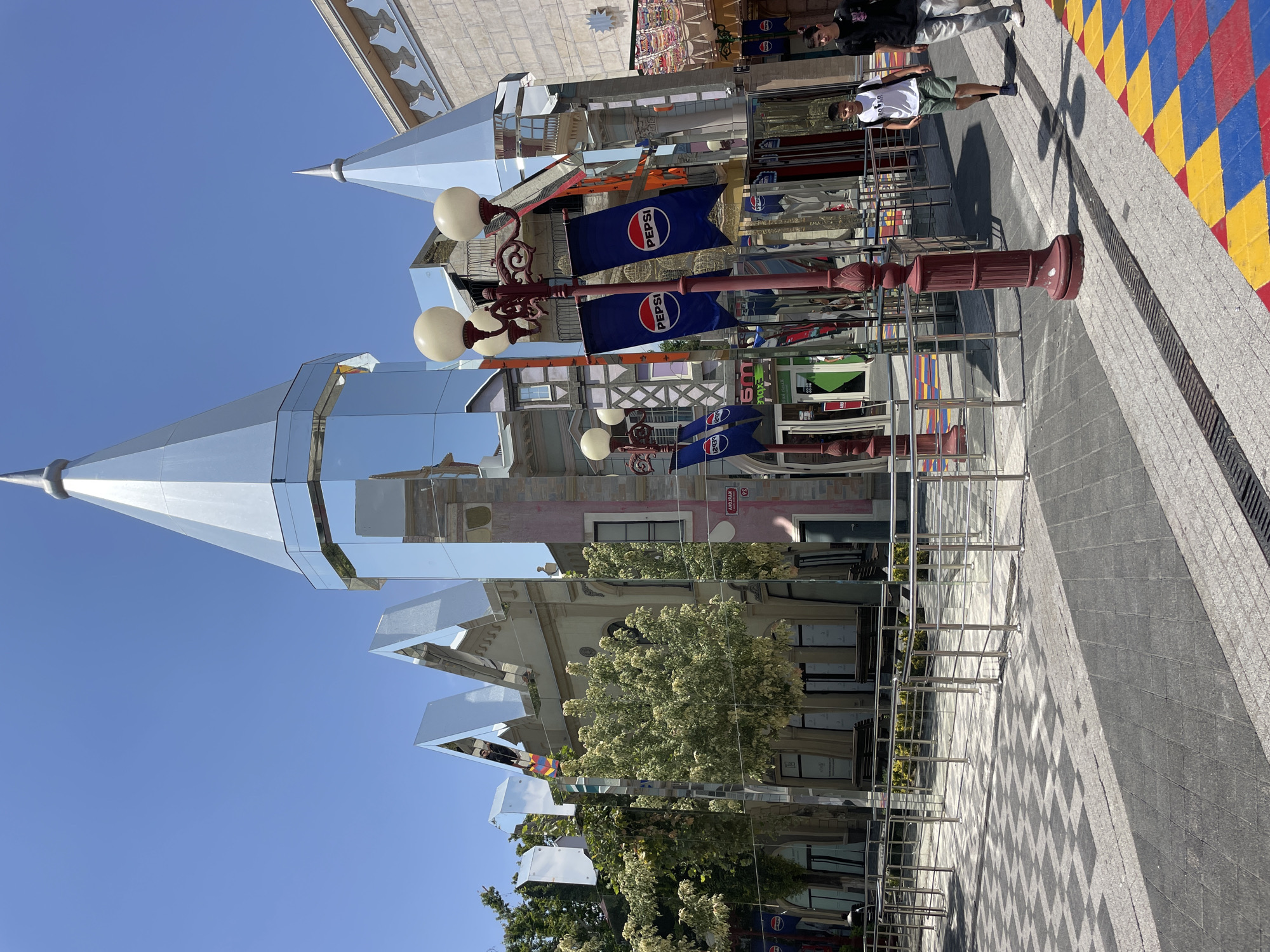 This is a photograph taken outdoors, likely on a sunny day given the clear blue sky and strong shadows. The image features a mirrored architectural structure that reflects the surrounding environment. Prominently visible are two lampposts with spherical lights and red decorative elements. Attached to these lampposts are blue flags with the Pepsi logo.
In the foreground, a checkered pavement in red, blue, and yellow is visible, suggesting a public walkway or plaza. There are two young men present. One is wearing a white t-shirt and green shorts, standing near the checkered pavement. The other, wearing a black t-shirt, is slightly further back. They appear to be casually walking or posing.
The mirrored building creates a disorienting and fragmented view of the streetscape. Reflections show buildings with windows and balconies, trees with lush green foliage, and more of the street and pavement. The overall scene suggests an urban environment, possibly a tourist area or a modern shopping district. The time of day appears to be daytime, with the sun high in the sky. The location cues, such as the checkered pavement and architectural style of the surrounding buildings reflected in the mirrored structure, combined with the specific Pepsi branding, hint at a contemporary setting.
