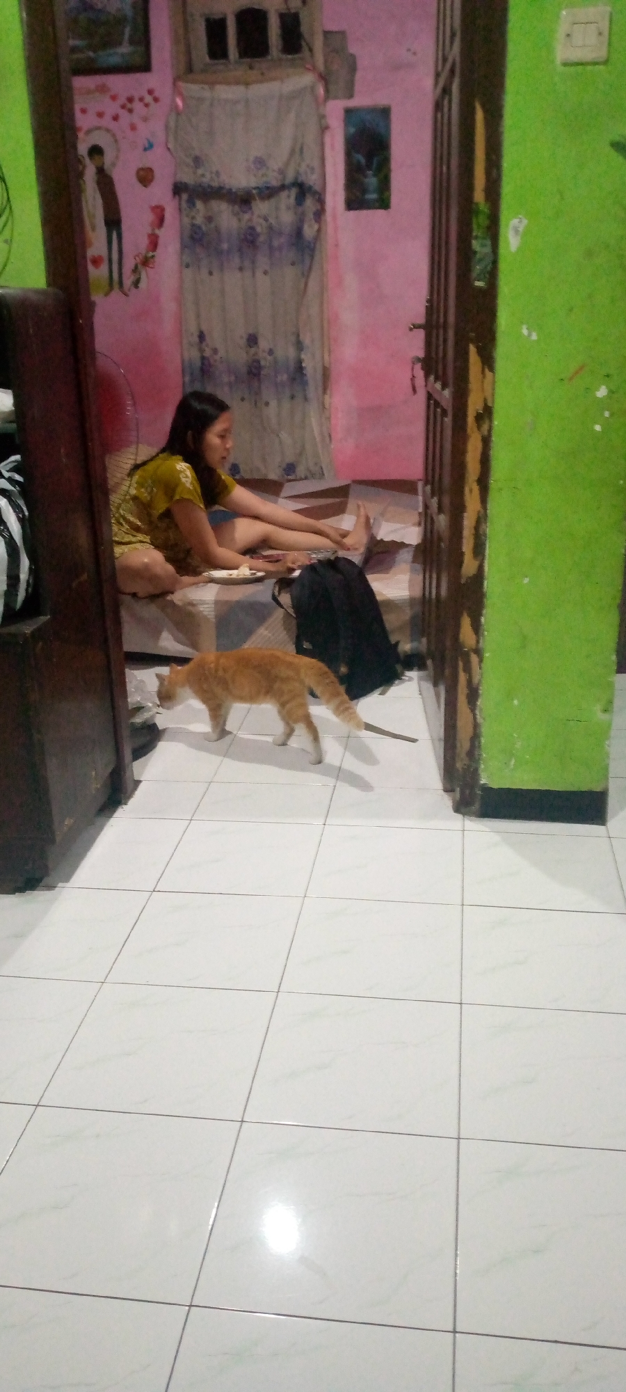 The image depicts an indoor scene within a residence in Sumberwono, Indonesia. A young woman with dark hair, wearing a yellow t-shirt and shorts, is seated on a bed with her legs extended. She appears to be holding a plate with food and is looking down. Beside her on the bed is a black backpack. On the floor in front of the bed, an orange tabby cat is walking away from the camera, its tail raised. The room has light-colored tiled flooring. The walls are painted pink and green, and a wooden door is visible on the right side of the frame. Decorative elements, including a framed picture and wall decals of hearts and a person, are present on the pink wall. A white electrical outlet is mounted on the green wall.