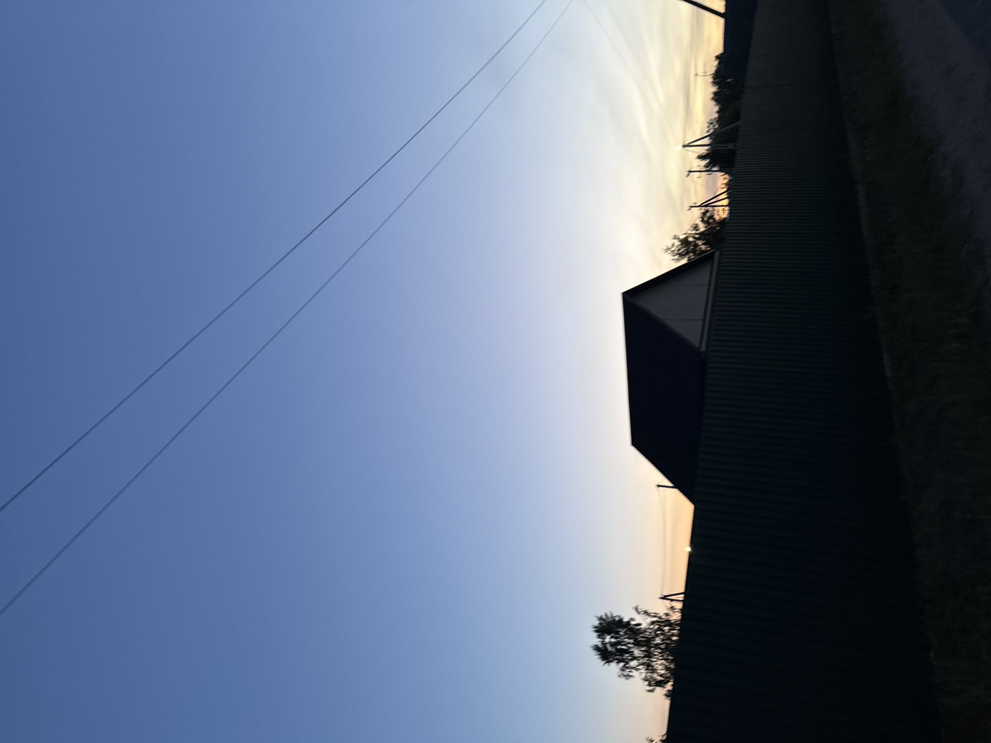 The image, when correctly oriented, presents a vast sky scene at either sunrise or sunset over a rural or semi-rural landscape. The sky dominates the frame, displaying a gradient from a clear, deep blue in the upper and left portions to a lighter, yellowish-orange tone near the horizon on the right. Two thin, dark power lines stretch diagonally across the upper-left sky.
The foreground elements are rendered in silhouette, indicative of the low sun position. A dark, corrugated fence runs horizontally across the lower-right side of the image, bordering a dark strip of ground or low vegetation. Behind the fence, a silhouetted building, possibly a house or shed with a distinct pitched roof, is partially visible on the right. Scattered along the horizon, dark silhouettes of trees and bushes are visible, with some branches extending above the fence and building. In the far distance on the right, several faint, dark shapes suggest utility poles or other structures against the lighter sky.
The scene captures the peaceful transition of day, with no human activity or individuals visible. The weather appears clear. No visible text is present in the image. The setting suggests an open area with simple structures, aligning with the provided location context of Лабжанскі сельскі Савет, Belarus.
