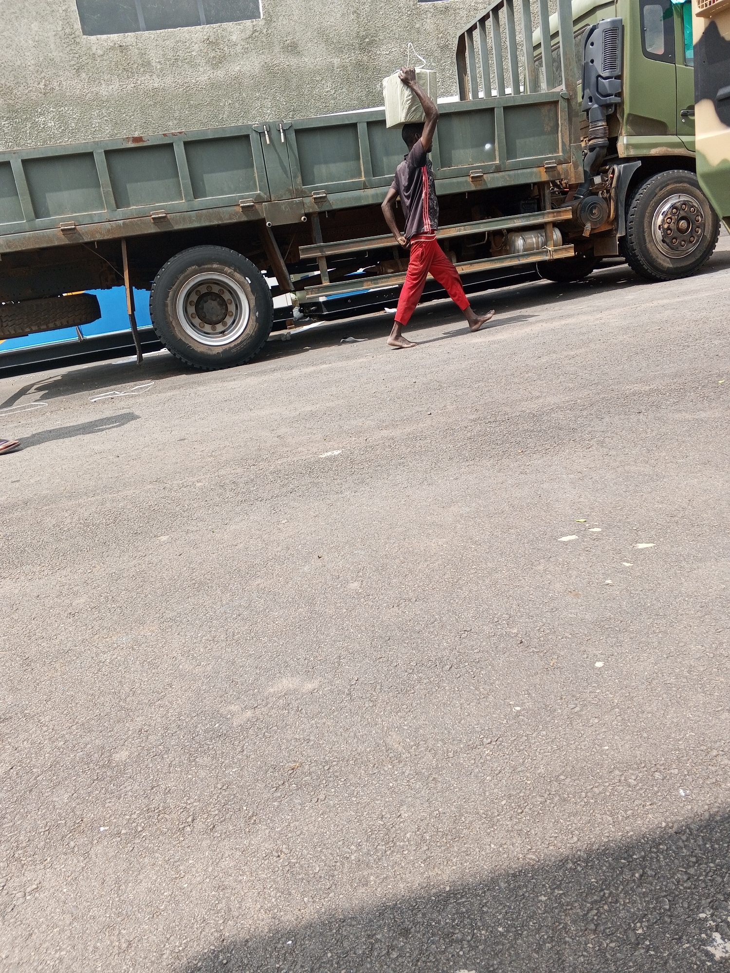 A man, barefoot and wearing a black and red shirt and red pants, carries a white object on his head while walking along a grey asphalt road next to a large green flatbed truck. The truck, with a visible wheel and part of its cabin, is parked parallel to the road and next to a building. The setting appears to be a street within a city, possibly Abuja, Nigeria.