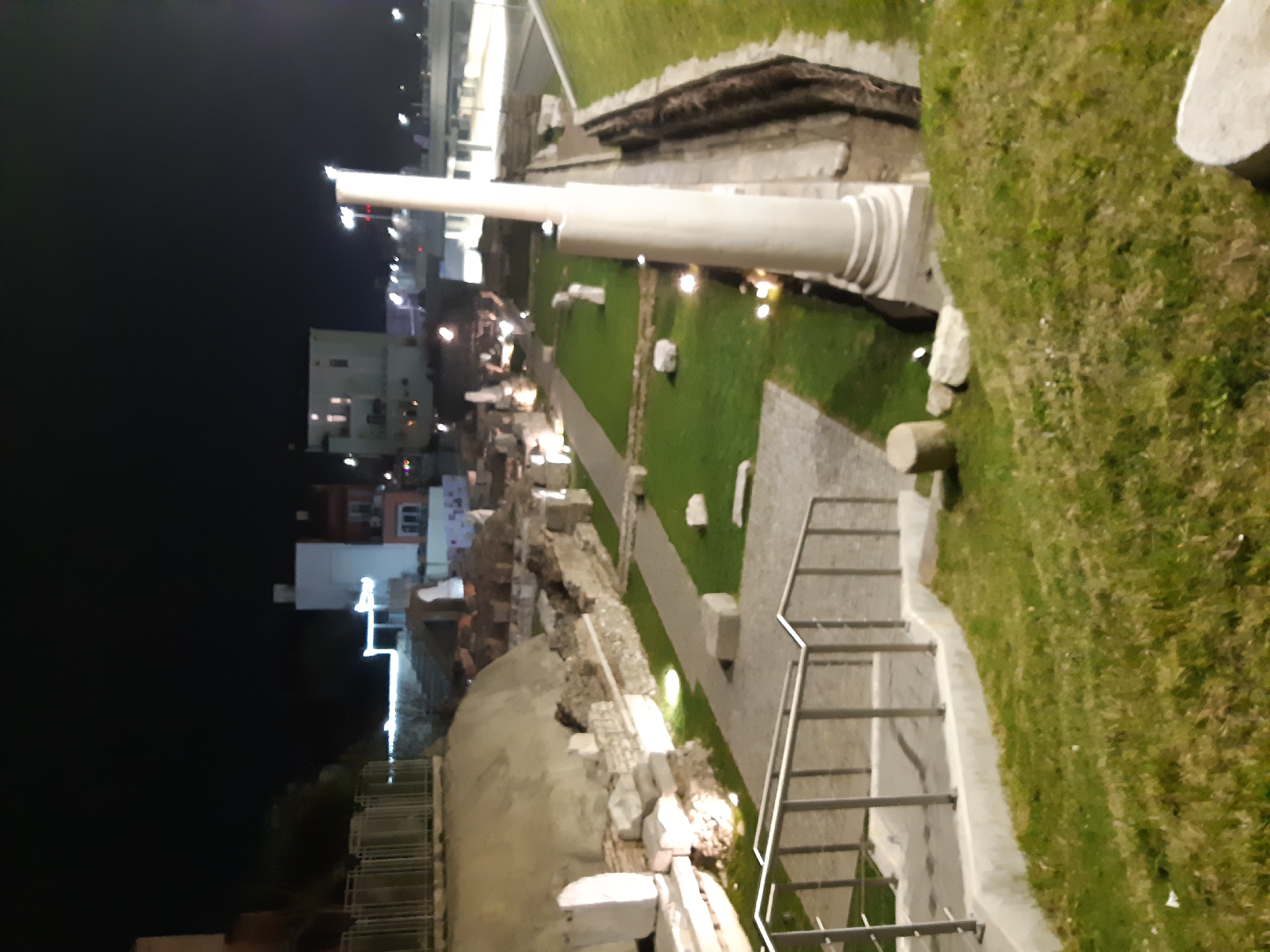 The image depicts the Roman Forum in Plovdiv, Bulgaria, at night. The ruins are illuminated, including a reconstructed column. A walkway leads through the site, with handrails visible in the foreground. Buildings are visible in the background beyond the ruins. Green grassy areas surround the stone structures. No people are visible in the immediate vicinity.