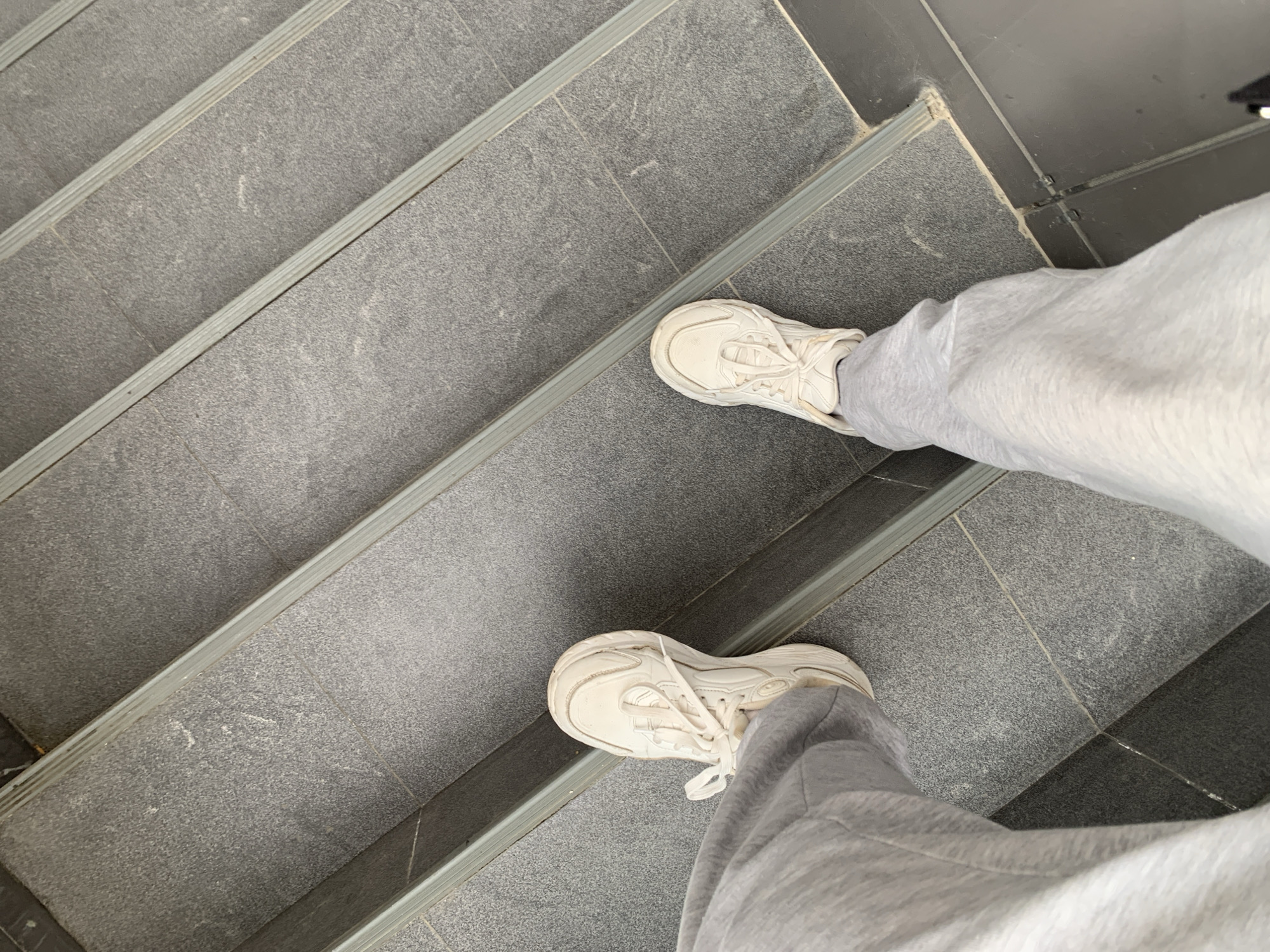An overhead, slightly angled shot captures the lower body of a person standing on a staircase. The individual is wearing light grey sweatpants or joggers and white, somewhat chunky athletic sneakers. Their right foot is positioned on a higher step, while their left foot rests on a lower one, suggesting a moment during ascent or descent.
The staircase is constructed from dark grey, speckled stone or concrete tiles. Each step features a lighter grey, ridged anti-slip strip along its edge. Portions of a darker, possibly black, tiled floor are visible in the top right corner and bottom left, indicating adjacent flooring. The lighting is soft and even, without strong shadows, making the time of day or specific weather conditions difficult to ascertain. There are no discernible emotions or visible text. The setting is a staircase in Stavropol, Russia.