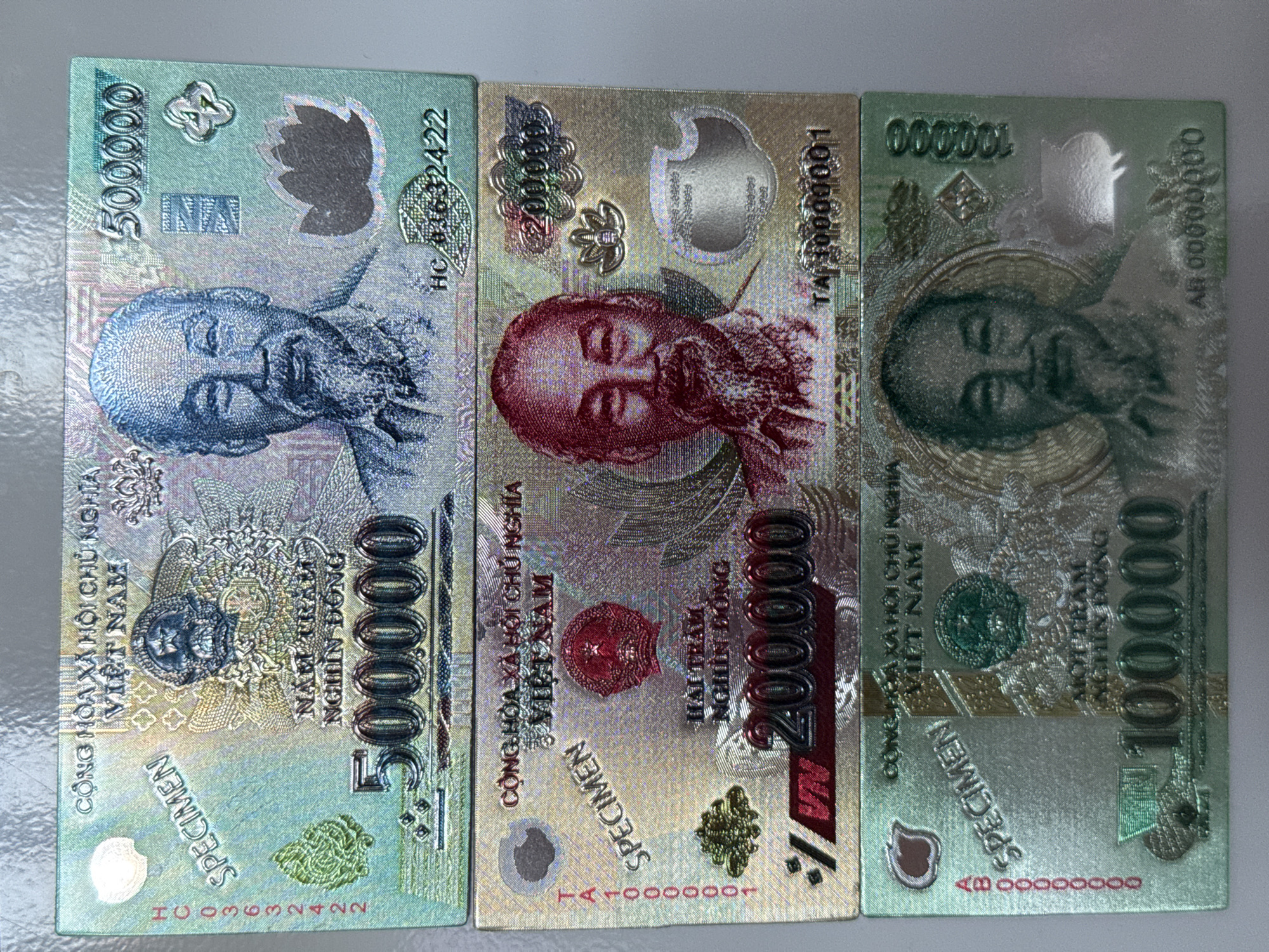 The image displays three Vietnamese banknotes, stacked vertically. The top banknote is green and features a portrait of Ho Chi Minh, along with the denomination "500000". The middle banknote is gold and features a portrait of Ho Chi Minh with the denomination "200000". The bottom banknote is also green and features a portrait of Ho Chi Minh with the denomination "100000". All banknotes appear to be in good condition with visible text and designs. The image does not provide any information about the location of these banknotes within the city of Izhevsk, Russia, nor does it depict any people, actions, or interactions. The background appears to be a textured surface, possibly a table or countertop.