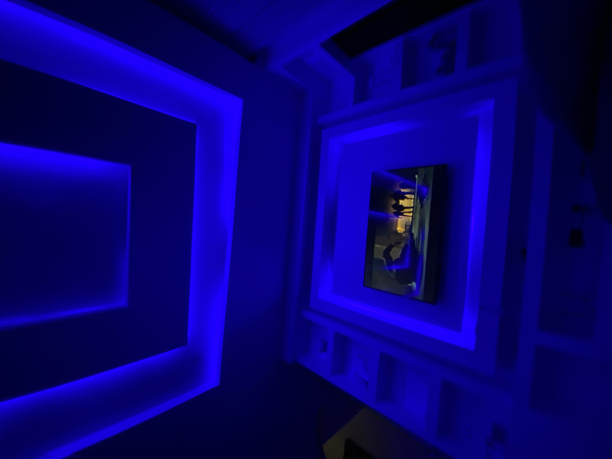 The image depicts an interior space with a modern design, possibly located in Lagos, Nigeria. The dominant feature is a series of square or rectangular recesses in the ceiling or wall, outlined with bright blue LED lighting, creating a striking visual effect. A television screen is mounted within one of these recesses, displaying what appears to be a scene with figures of people. The surrounding architecture consists of stepped, geometric elements also illuminated in blue. The lighting suggests a nightlife or entertainment setting. There are no people visible other than the images on the television. The atmosphere is modern and design-focused.