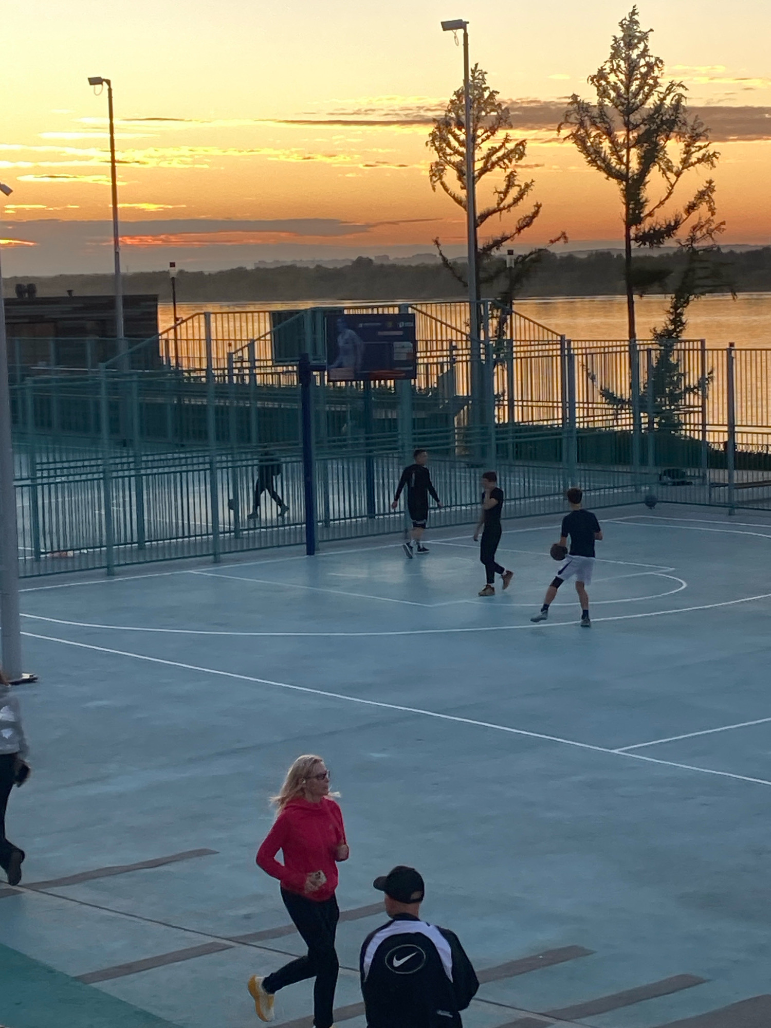 The image captures a basketball court at sunset, likely located at the embankment of the Kama River in Perm, Russia, given the presence of the river and the characteristic sky. The sky is ablaze with warm hues of orange and yellow, with scattered clouds. Two silhouetted trees with sparse foliage stand tall against the horizon.
On the basketball court, four individuals are visible. Three young men are engaged in a basketball game. One is dribbling a ball, another is standing and observing, and a third is positioned further back on the court. A woman in a bright pink hoodie and black leggings is jogging on the perimeter of the court, wearing glasses. In the foreground, a man wearing a black Nike jacket and cap is seated on bleachers, facing away from the viewer.
The basketball court itself is a light blue surface marked with white lines. It is enclosed by a green metal fence, behind which the river and the distant shoreline are visible. A light pole with a lamp stands to the left of the frame. The overall impression is a serene evening scene with recreational activities taking place against a backdrop of a beautiful sunset.