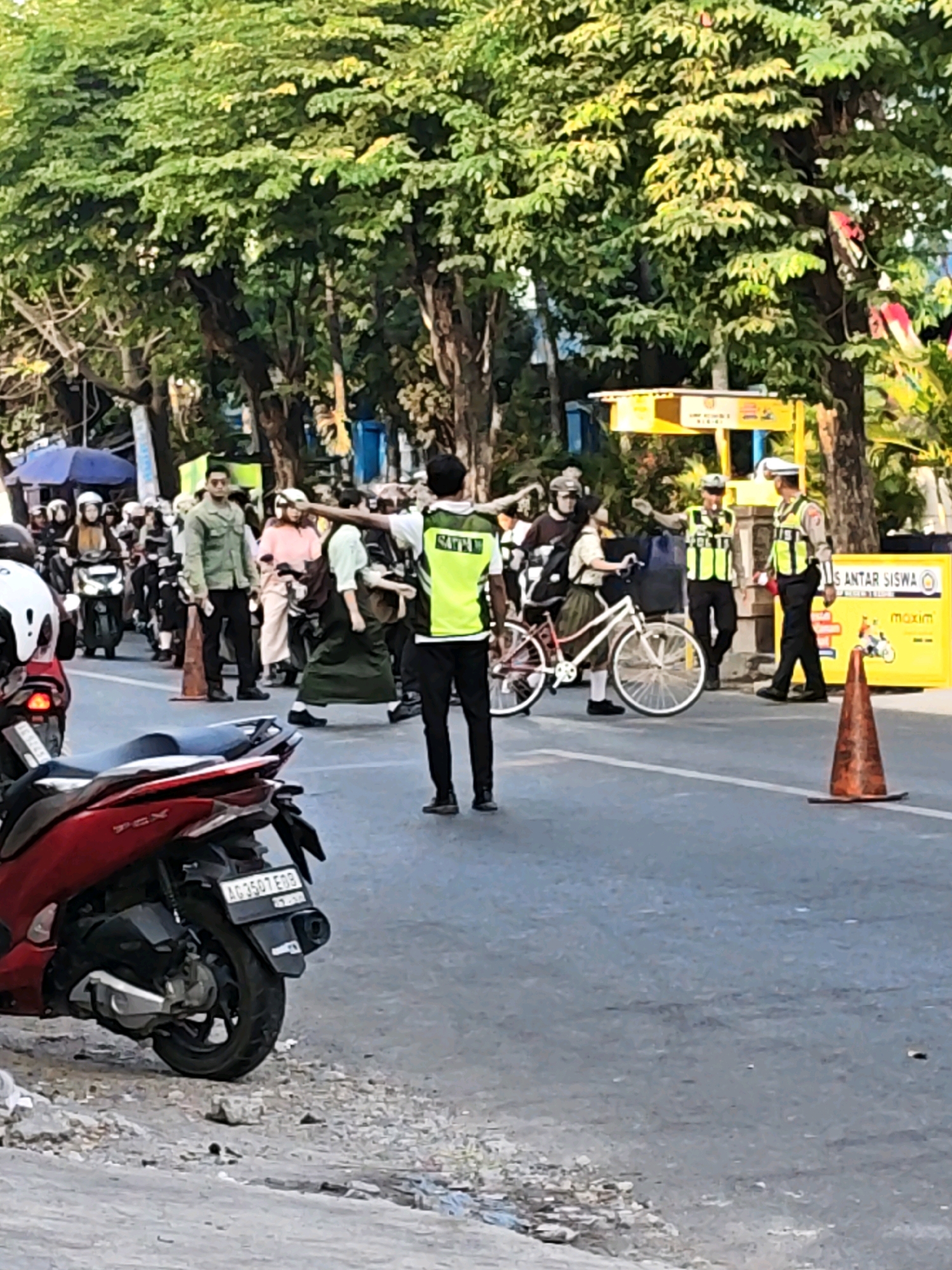 In the city of Kediri, Indonesia, traffic is being managed near a road sign that reads "SANTAR SISWA" and "Maxim." A person in a reflective vest, likely a traffic director or officer, is gesturing with their left arm, directing traffic. Several individuals are on motorcycles, with some appearing to be students in uniform. One student is pushing a bicycle. Orange traffic cones are visible on the road. The scene appears to be a busy street with trees lining the sides.