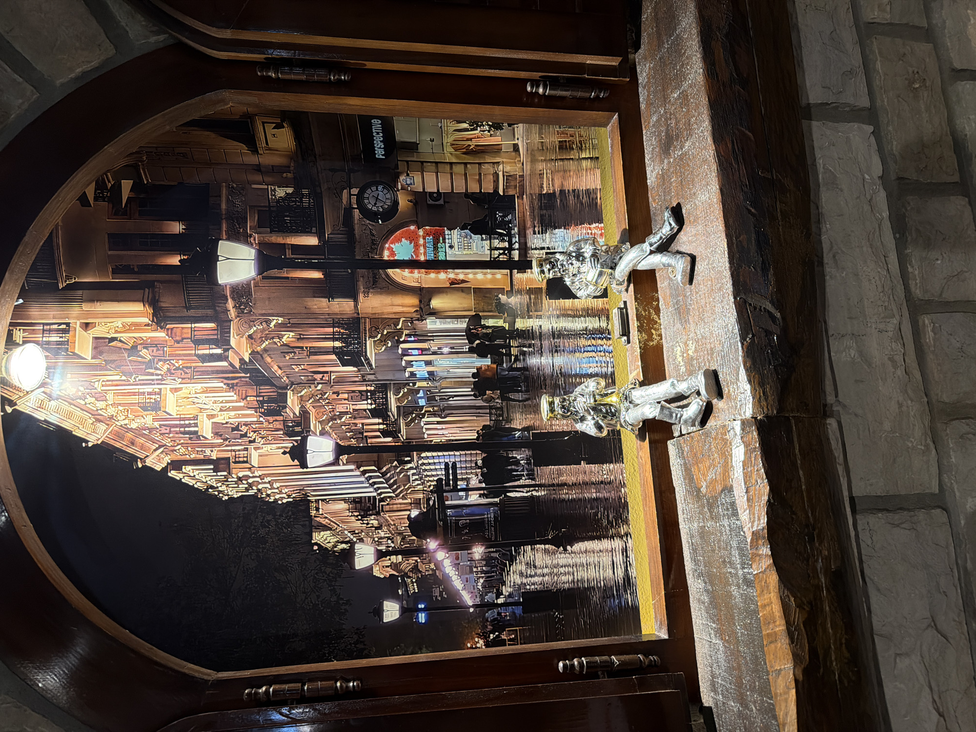 The image shows a view through an arched wooden window frame onto a nighttime city street scene. The street is wet, reflecting the lights from buildings and streetlamps. People are visible walking along the sidewalk, some with umbrellas. Two small, silver-colored figurines are seated on the wooden windowsill in the foreground. One figurine is playing a trumpet, and the other is holding a drum. The background depicts buildings with ornate architecture, illuminated by numerous lights. The scene is set in the city of Kumbor, Montenegro, as indicated by the presence of "Perspective" signage on one of the storefronts, which is a known establishment in Kumbor.
