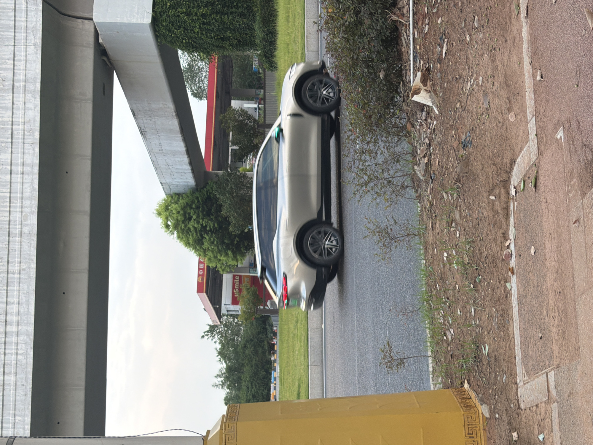 The image shows a silver SUV driving on a road in Jiangjin, China. The car is positioned in the center of the frame, moving from left to right. In the background, a red and white gas station with a "中国石油" (PetroChina) sign is visible, along with green trees and a cloudy sky. The foreground features a yellow pillar on the left side of the frame, a patch of dry dirt with sparse vegetation, and a section of sidewalk. Overhead, a concrete overpass stretches across the top of the image. The scene appears to be an urban or suburban environment.