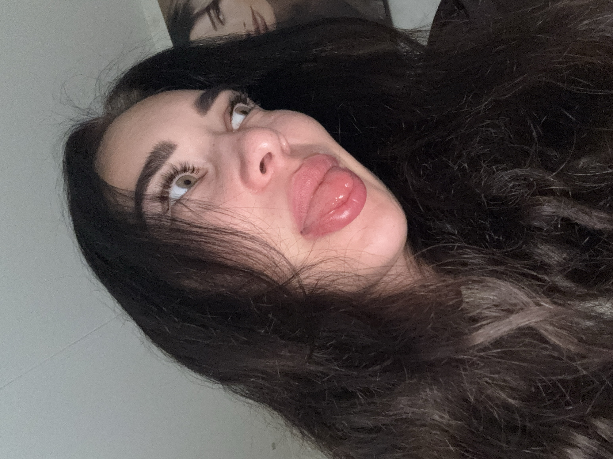 A close-up, angled portrait features a young woman with long, dark, wavy hair cascading around her face. Her head is tilted, and she gazes upwards and to the right side of the frame with light-colored eyes, possibly hazel or light brown. Her eyebrows are dark and well-defined, complemented by long, distinct eyelashes. Her full lips, which appear glossy, are slightly open, with her tongue extended to touch her upper lip, creating an expressive and somewhat playful or suggestive pose.
The background is a plain, light-colored surface, likely a wall, with hints of a faint, out-of-focus image of another face (possibly a drawing or poster) visible behind her head, partially obscured by her hair. The lighting is soft and even, suggesting an indoor setting without harsh shadows. No text is visible in the image.