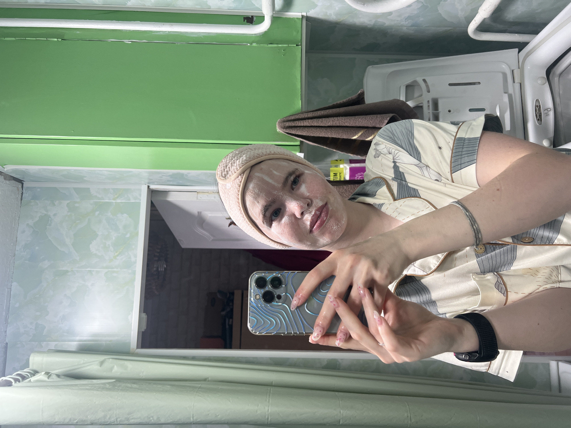 An indoor mirror selfie shows a young woman applying a white face mask. She has light-colored hair and eyes, and wears a beige terry cloth headband. Her face is partially covered with the white mask, applied in streaks. She is dressed in a light-colored, patterned pajama-style top. On her left wrist, she wears a black smartwatch, and a silver bracelet is visible on her right arm. Her fingernails are long and manicured with a clear or light pink polish and small, embedded glitter or designs.
She holds a smartphone, likely an iPhone with a triple-lens camera, which has a distinctive blue and silver wavy patterned case. The image is oriented sideways, appearing as if the subject is lying down.
The background, visible in the mirror reflection, depicts what appears to be a bathroom or utility area in a home in Chaikovsky, Russia. Above her reflection is a vibrant green painted surface, possibly a cabinet or shelf, with white piping running along the top. To her right, a white washing machine or similar appliance is visible with its lid open. A folded brown towel hangs nearby. On a shelf behind her, various bottles and containers are stacked, with partially legible labels including "2M" and fragments like "no" and "EL". The walls feature a light green and white patterned surface, resembling marble or abstract wallpaper/tiles. A light green sheet or curtain is visible in the lower left foreground. The overall setting suggests a private and relaxed environment.