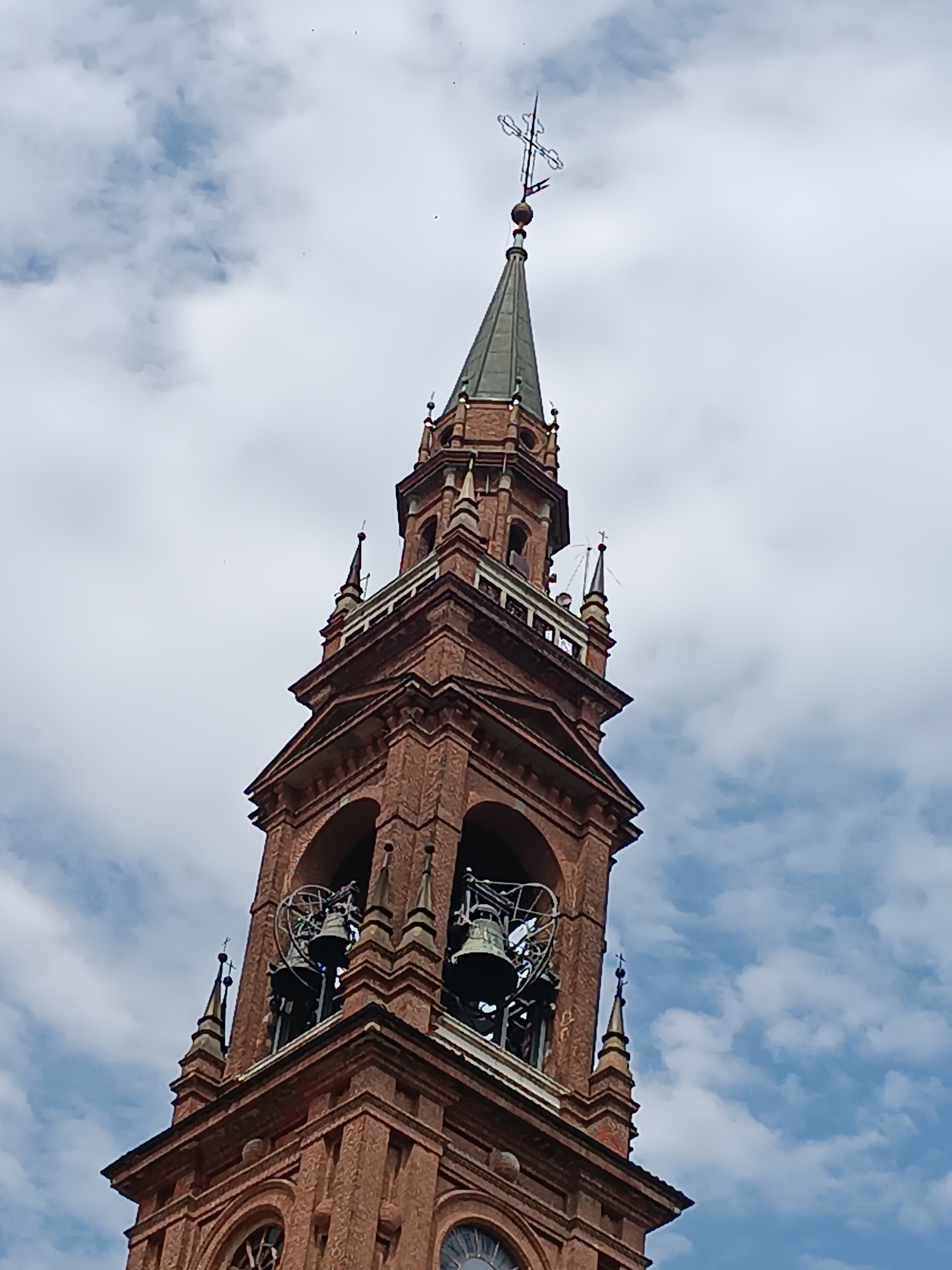 The image shows a close-up, low-angle view of a red brick bell tower, likely part of a church or cathedral. The tower has multiple levels with arched openings where bells are visible. The top of the tower is topped with a spire and a decorative cross. The sky in the background is partly cloudy. The architectural style suggests it is located in Europe, possibly Italy. Based on the specific architectural features, this is likely the bell tower of the Duomo of Casalmaggiore, Italy.
