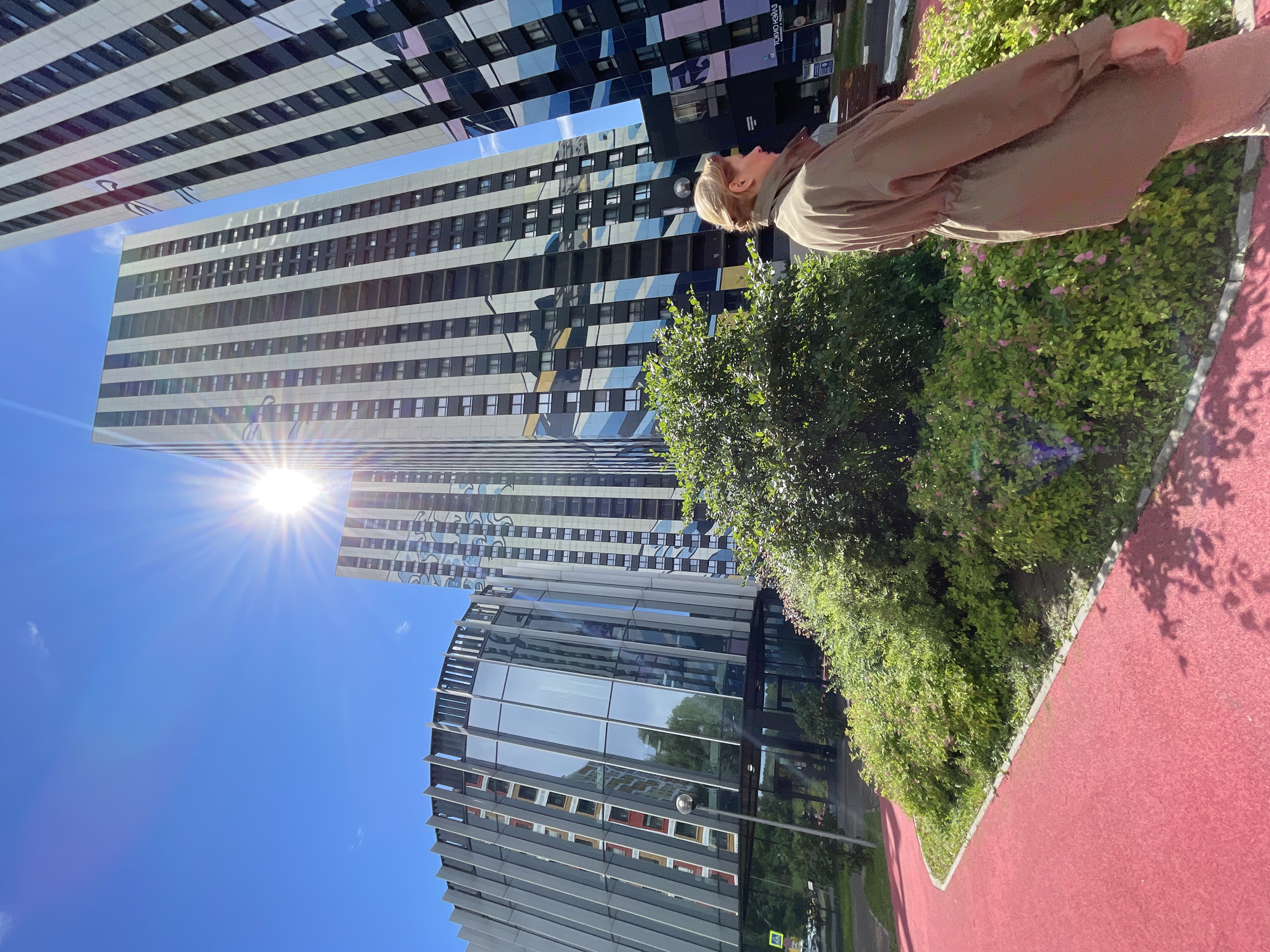 A woman stands on a red walking path in front of a lush green bush. Behind her, several modern high-rise buildings with distinctive geometric facades pierce a clear blue sky, with the sun flaring brightly in the upper left corner. The architecture includes a curved building with a glass facade and vertical metallic fins, and other towers clad in alternating black and white vertical stripes or colorful geometric patterns. The scene appears to be set in a newly developed urban area.