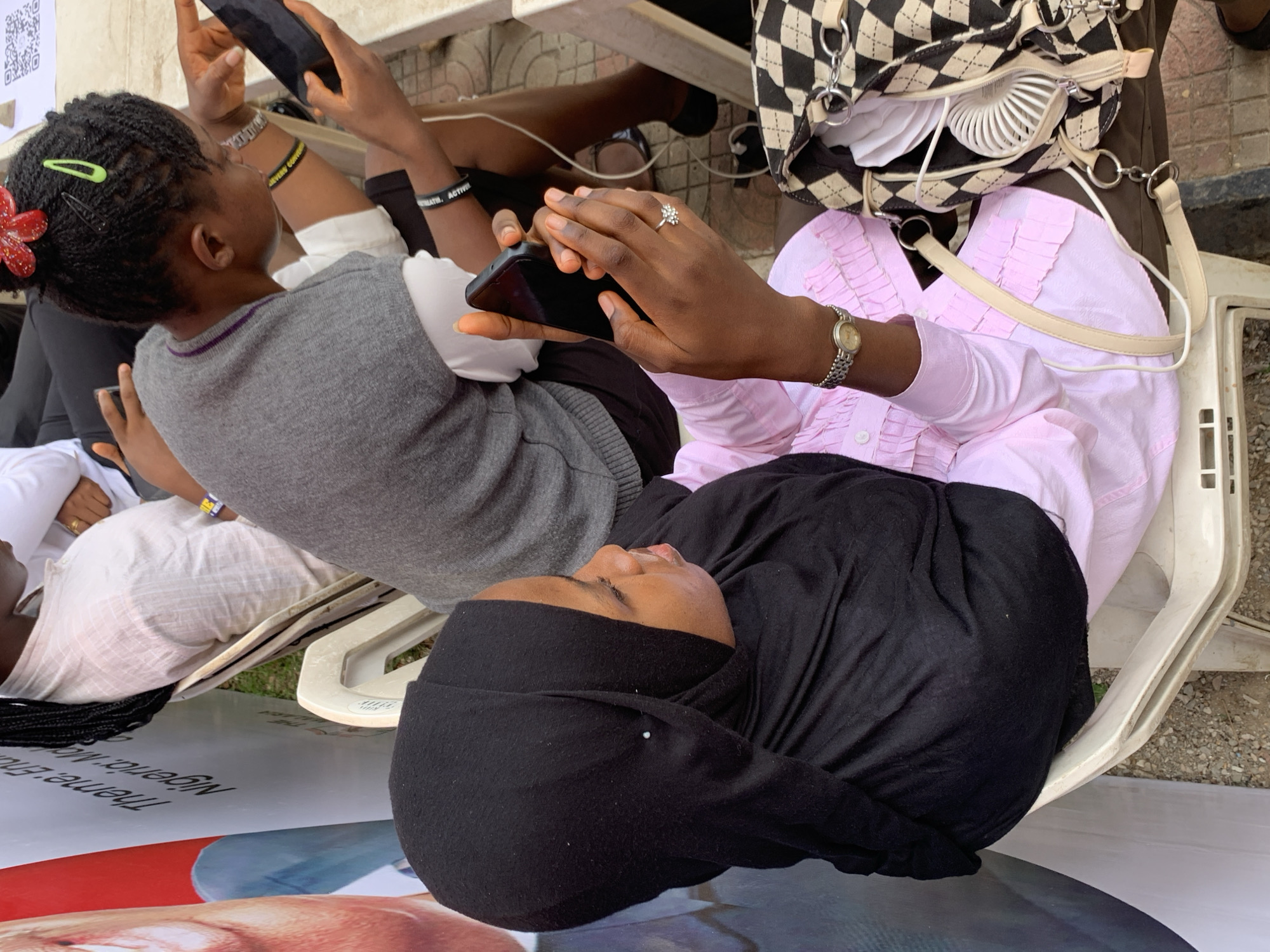 Captured from an elevated perspective, the scene depicts several individuals seated outdoors. One person wears a black hijab and a pink, long-sleeved top. Another individual holds a smartphone and has braided hair adorned with a red flower and green clips. A purse featuring a black and white checkered pattern rests on one of the chairs. The people are seated on white plastic chairs. The scene appears to be in Ilorin, Nigeria, as suggested by the text, "Nigeria: The Theme" on a graphic visible on the ground.