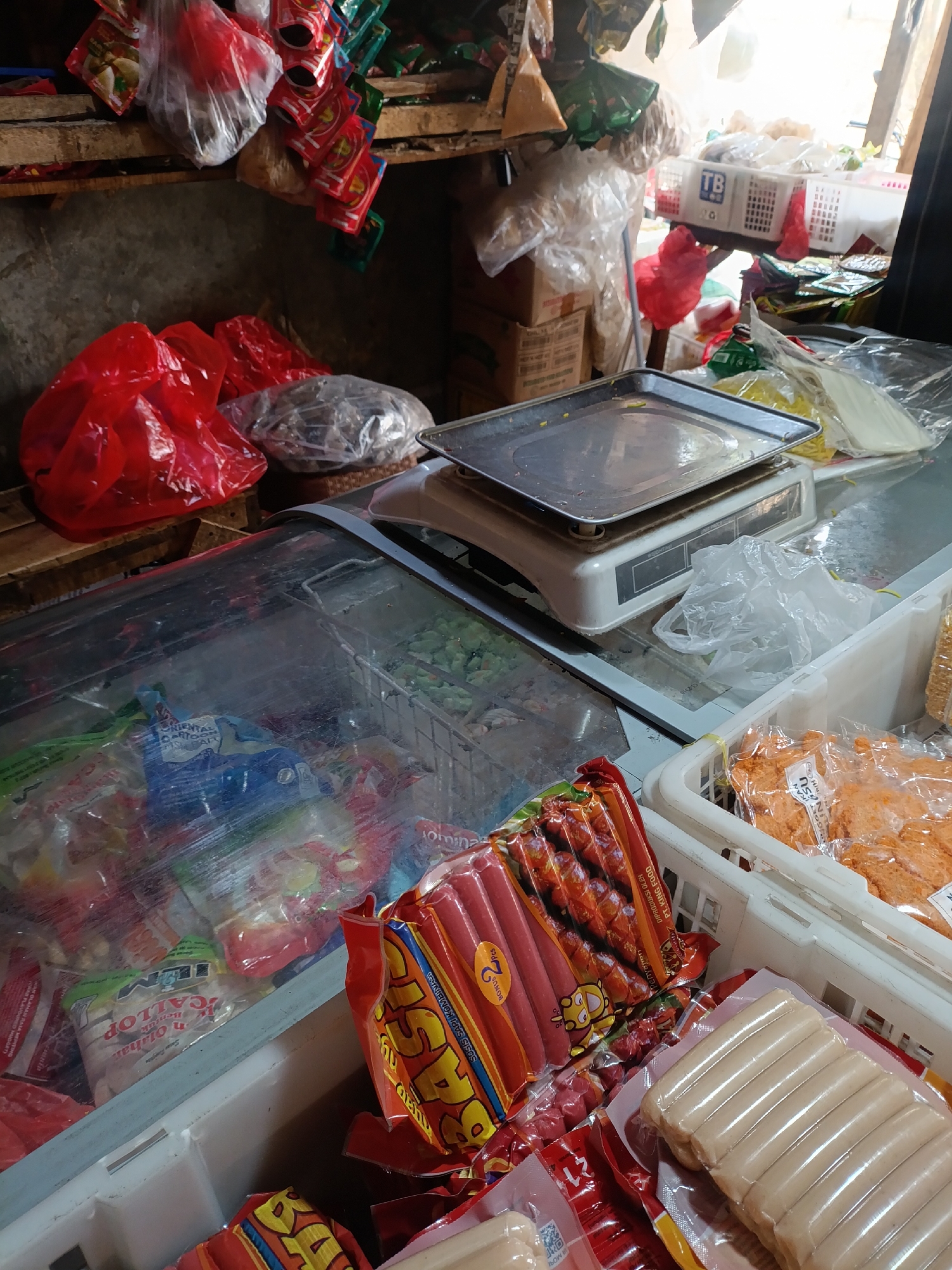 The image depicts the interior of a small, cluttered shop, likely a warung or general store, in Cariu, Indonesia. The scene is dominated by shelves packed with various food items, packaged goods, and storage containers. In the foreground, a commercial freezer displays an assortment of frozen food products, including what appear to be sausages and seafood. To the right of the freezer, a white plastic basket holds an orange, breaded food item. A digital scale sits on the counter, suggesting a point of sale. The overall impression is one of a busy, functional marketplace stall.